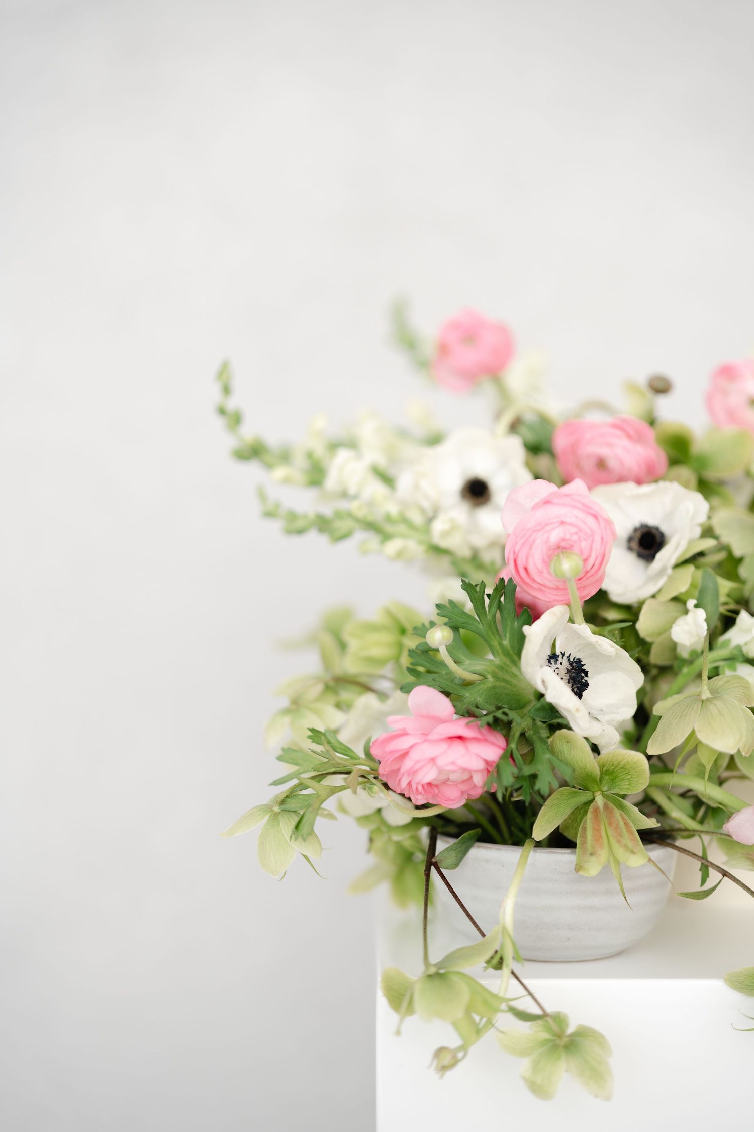 A white ceramic vase with pink ranunculus, white anemones, and green foliage arranged on a white surface.
