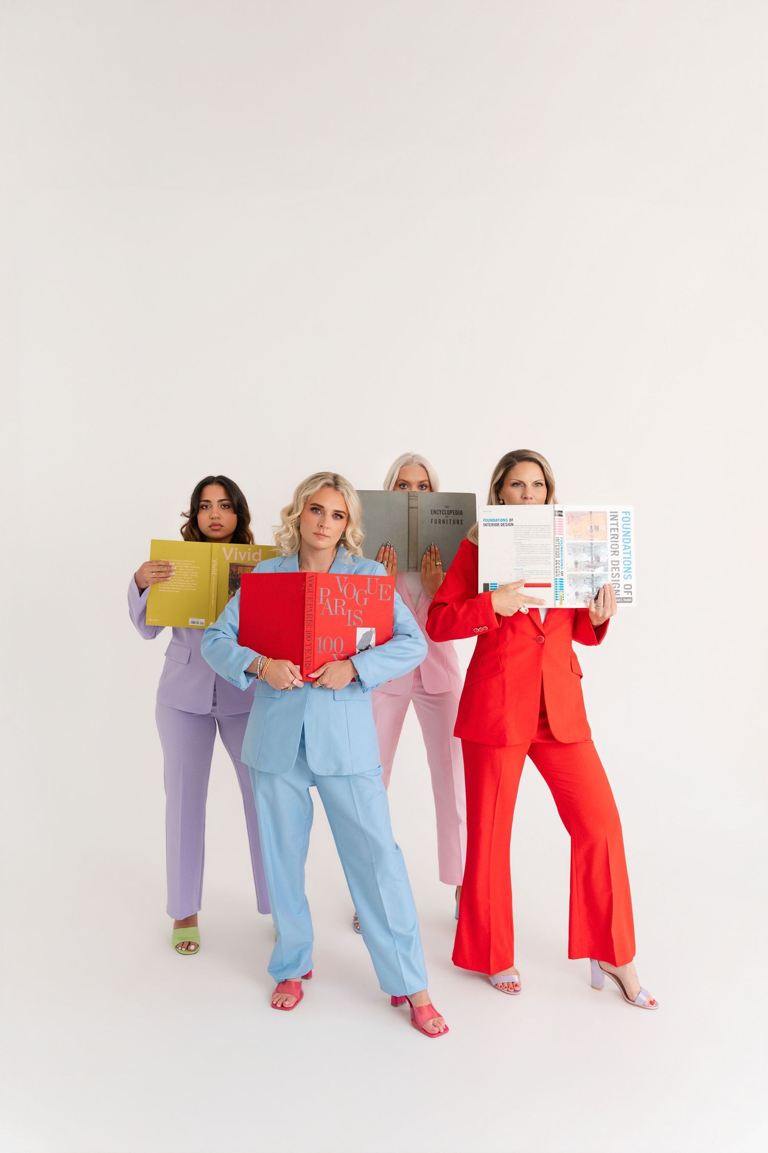Four women in colorful suits holding books pose against a white background in a vibrant fashion editorial.
