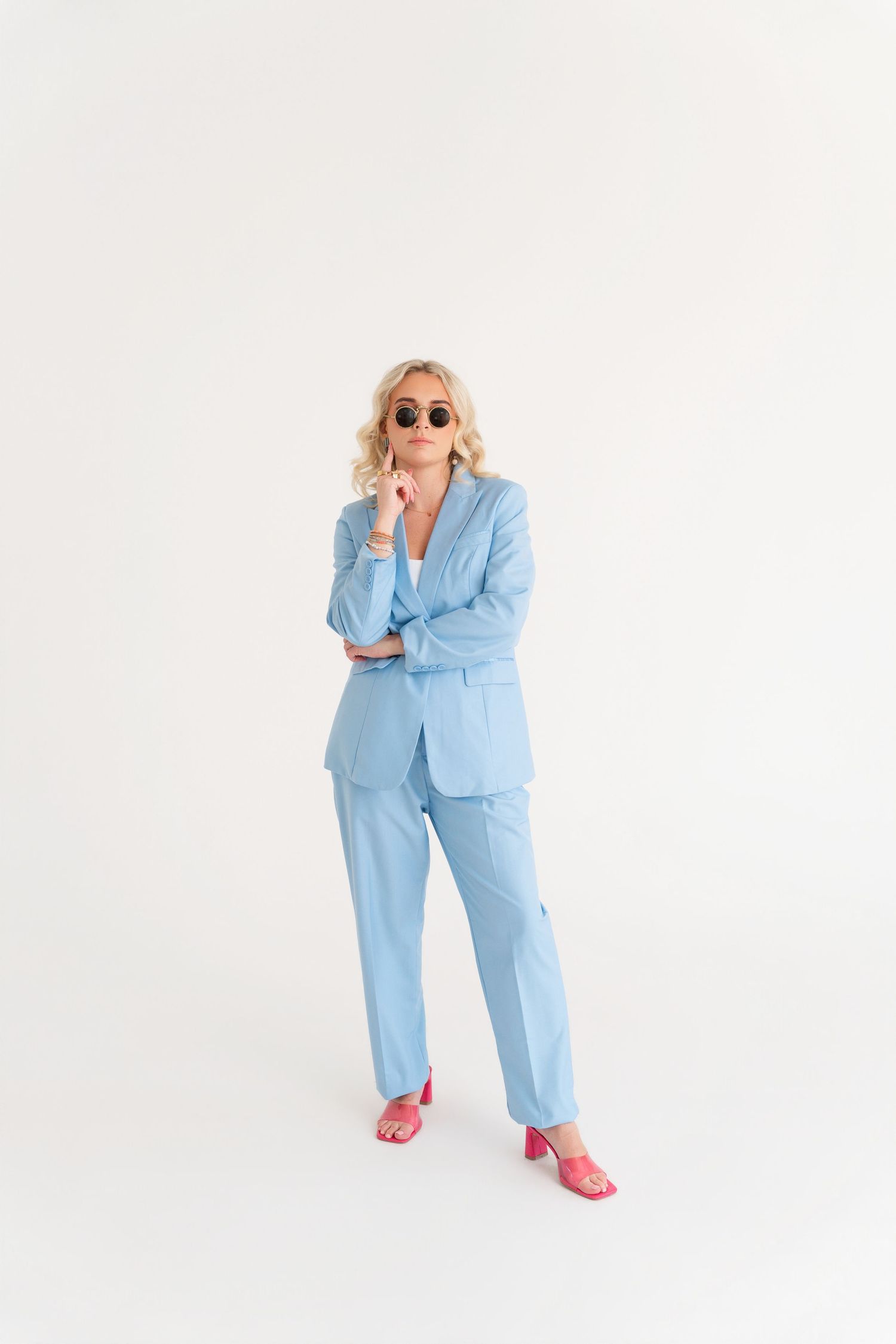 Woman in light blue pantsuit and pink heeled sandals wearing sunglasses poses confidently on white background.