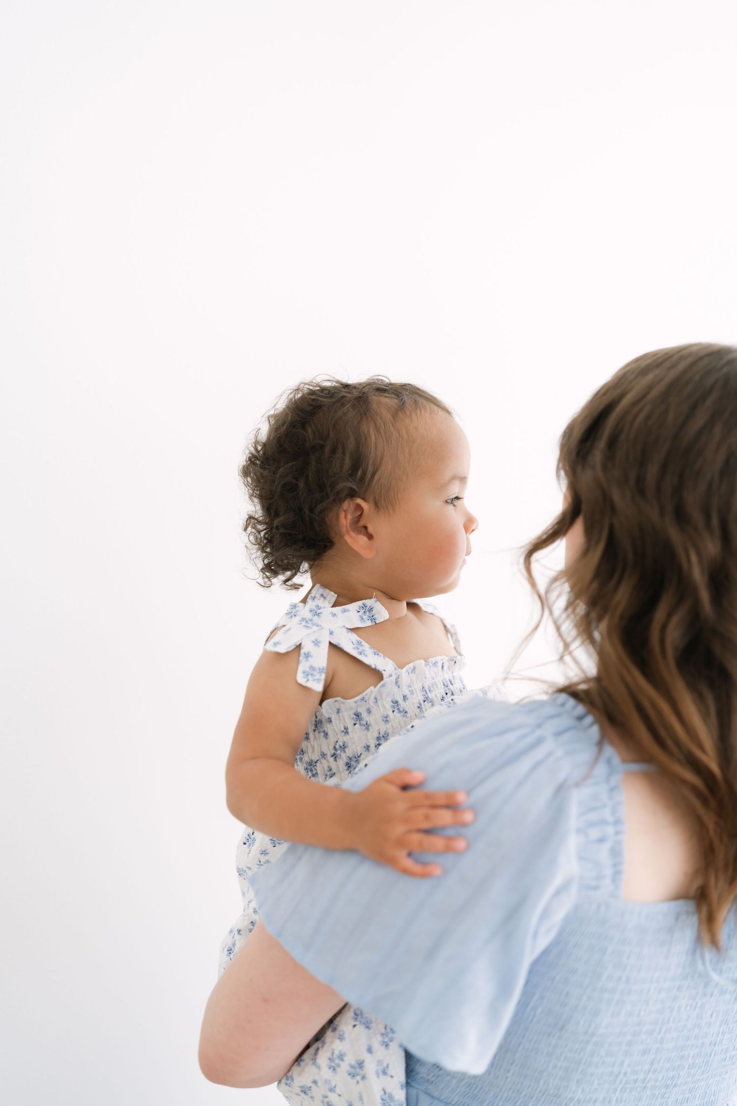 A mother in a blue top holds her curly-haired baby girl wearing a floral dress with a bow.