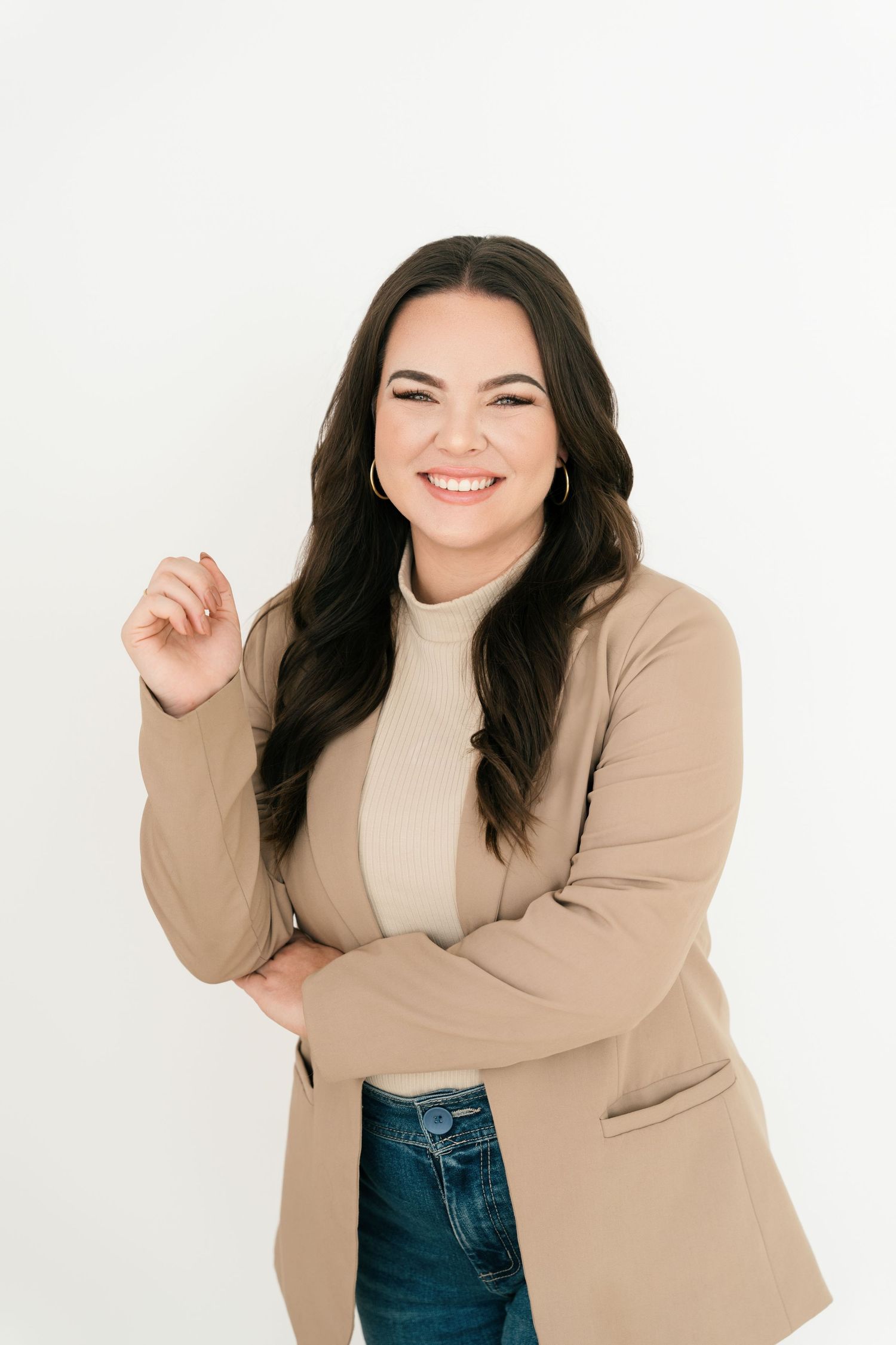 Smiling woman with dark hair wearing a tan blazer and turtleneck with jeans, posing confidently on white background.