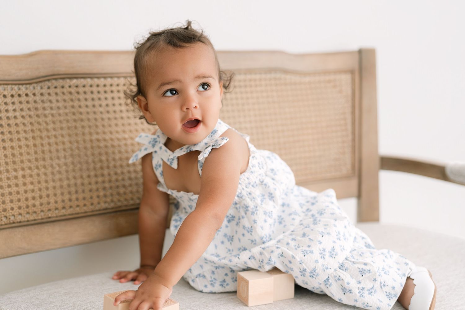 A baby girl in a floral romper sits on a bed, looking up curiously with a wooden block toy nearby.