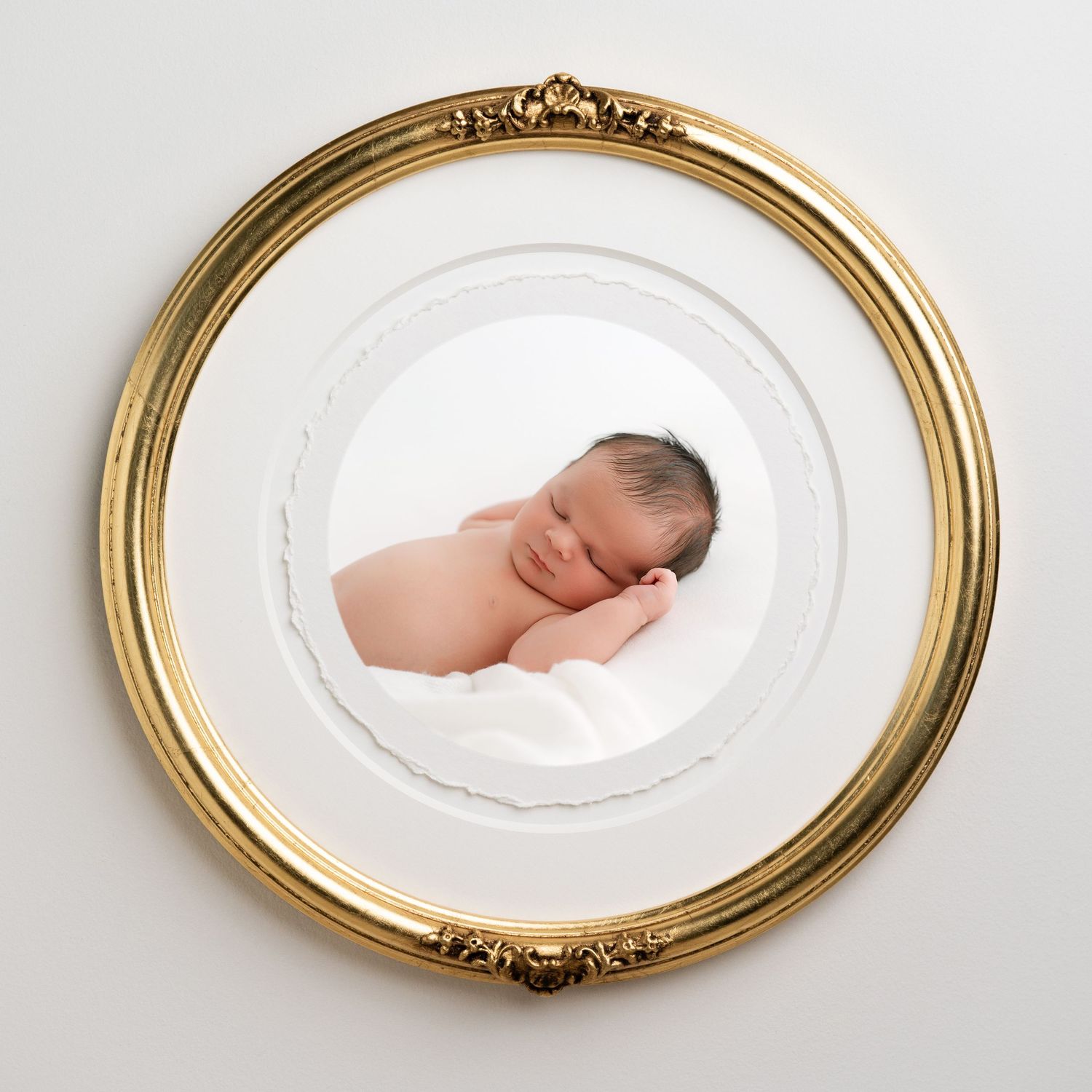 Sleeping newborn baby in a white wrap, displayed in an ornate circular gold frame on a white wall.