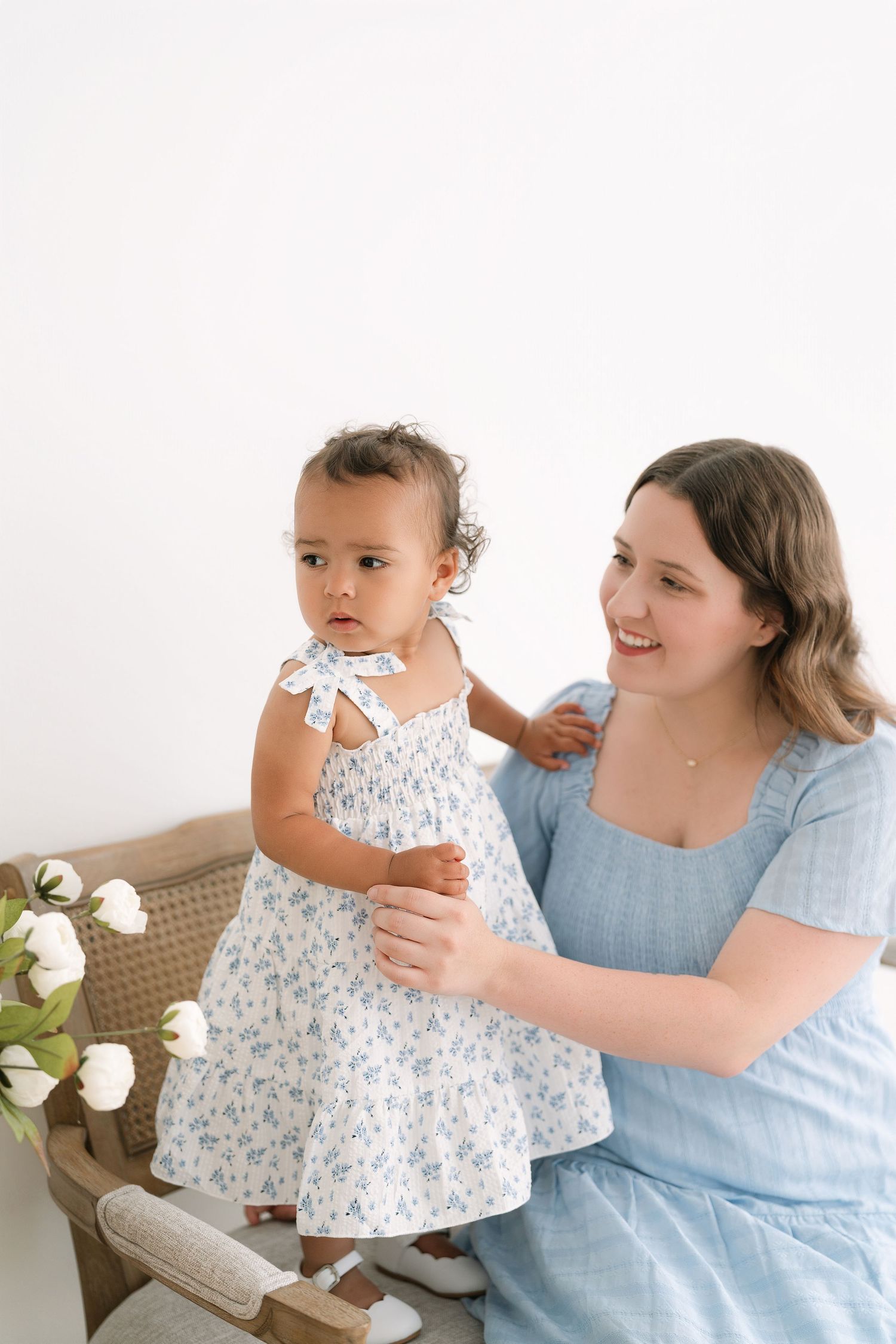 A smiling mother in a blue dress holds her toddler daughter in a white floral dress during a studio photo session.