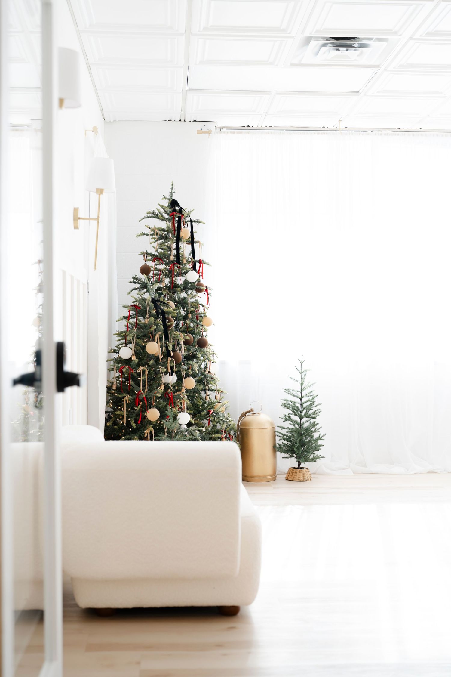 Bright white living room with decorated Christmas tree, small pine tree, and white sofa for a minimalist holiday decor.