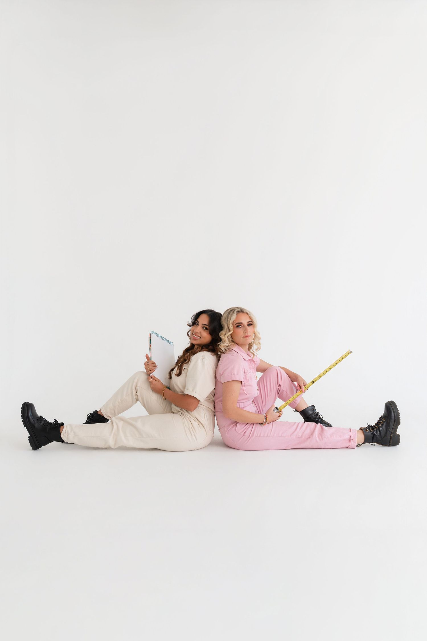 Two women in matching outfits sit back-to-back on white floor, one holding tablet, other holding yellow ruler.