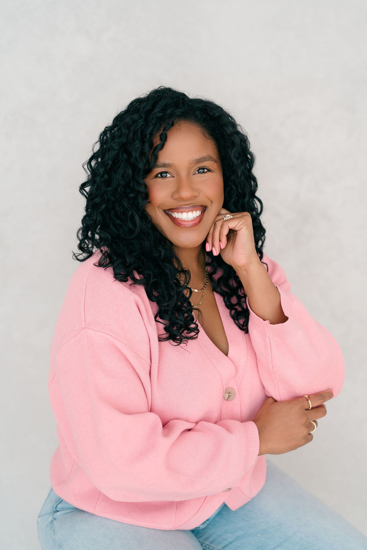 Smiling woman with curly hair wearing a pink jacket and jeans, posing confidently against a light gray background.