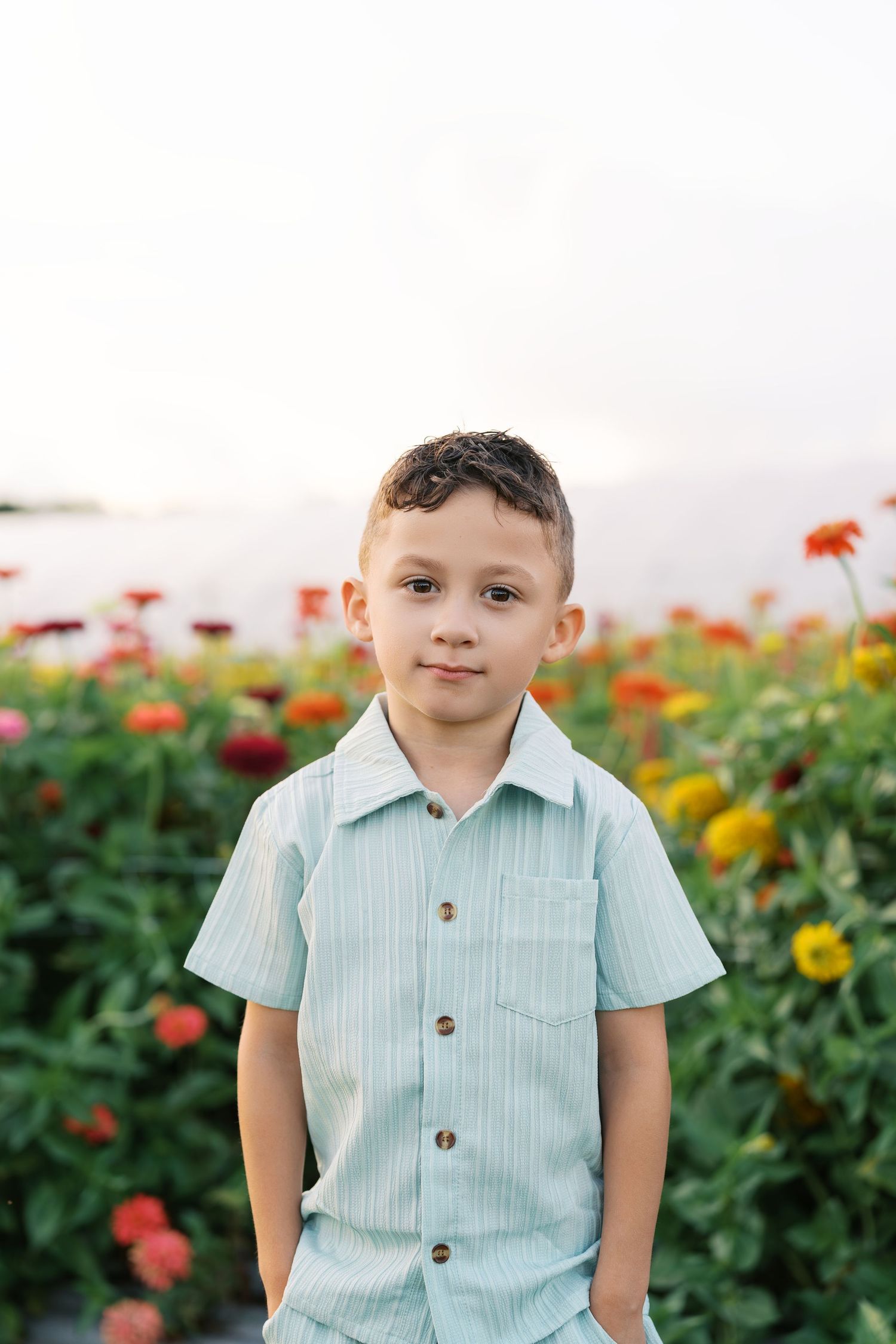 Young boy in light blue striped shirt standing in front of colorful flower garden, smiling at camera.