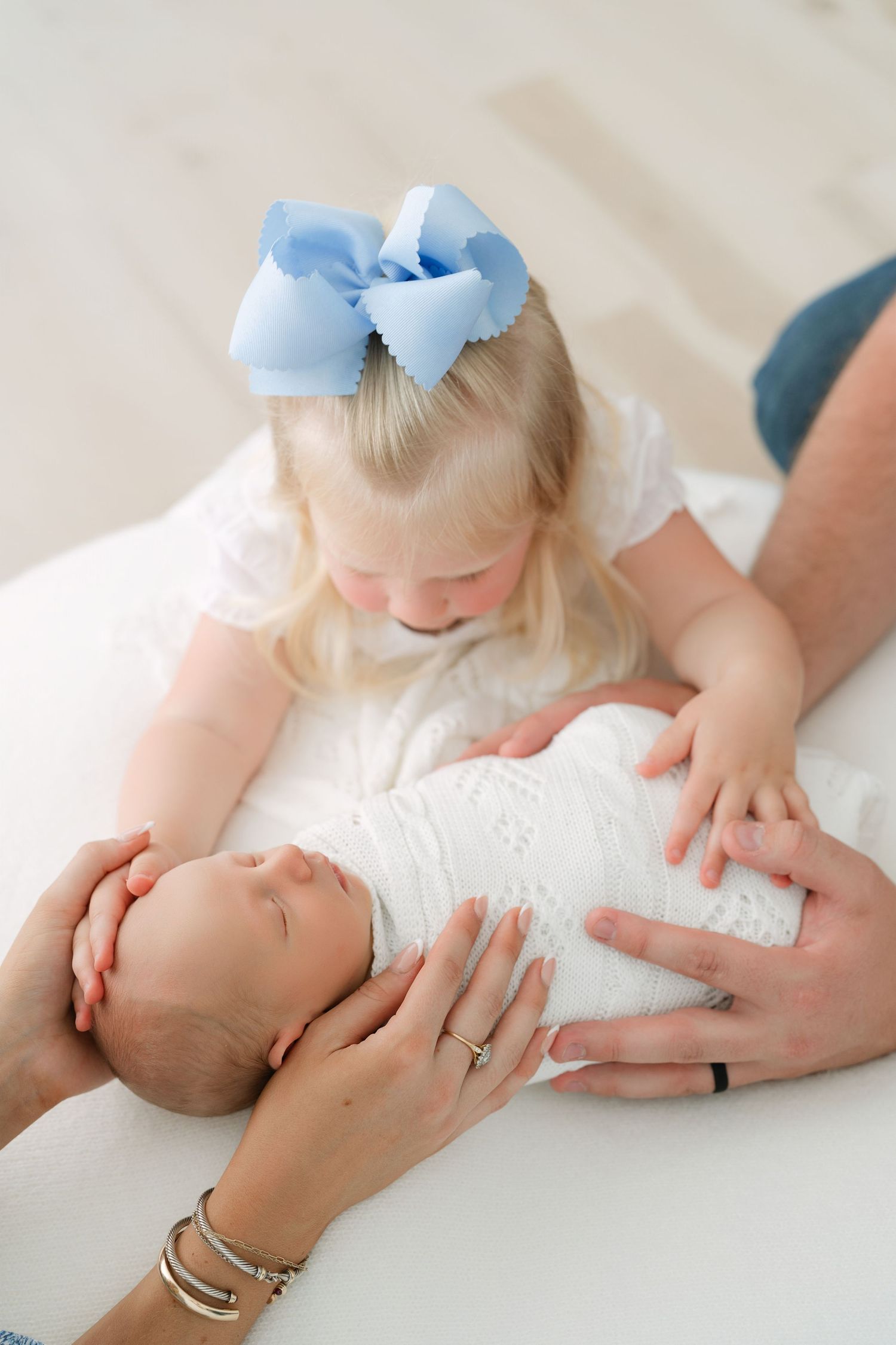 A toddler girl with a blue bow looks lovingly at a swaddled newborn baby held by parents hands.