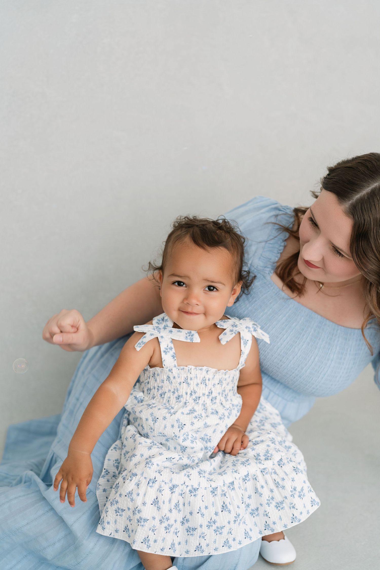 A mother in a blue dress lovingly gazes at her smiling baby girl in a white floral dress during a studio photo session.