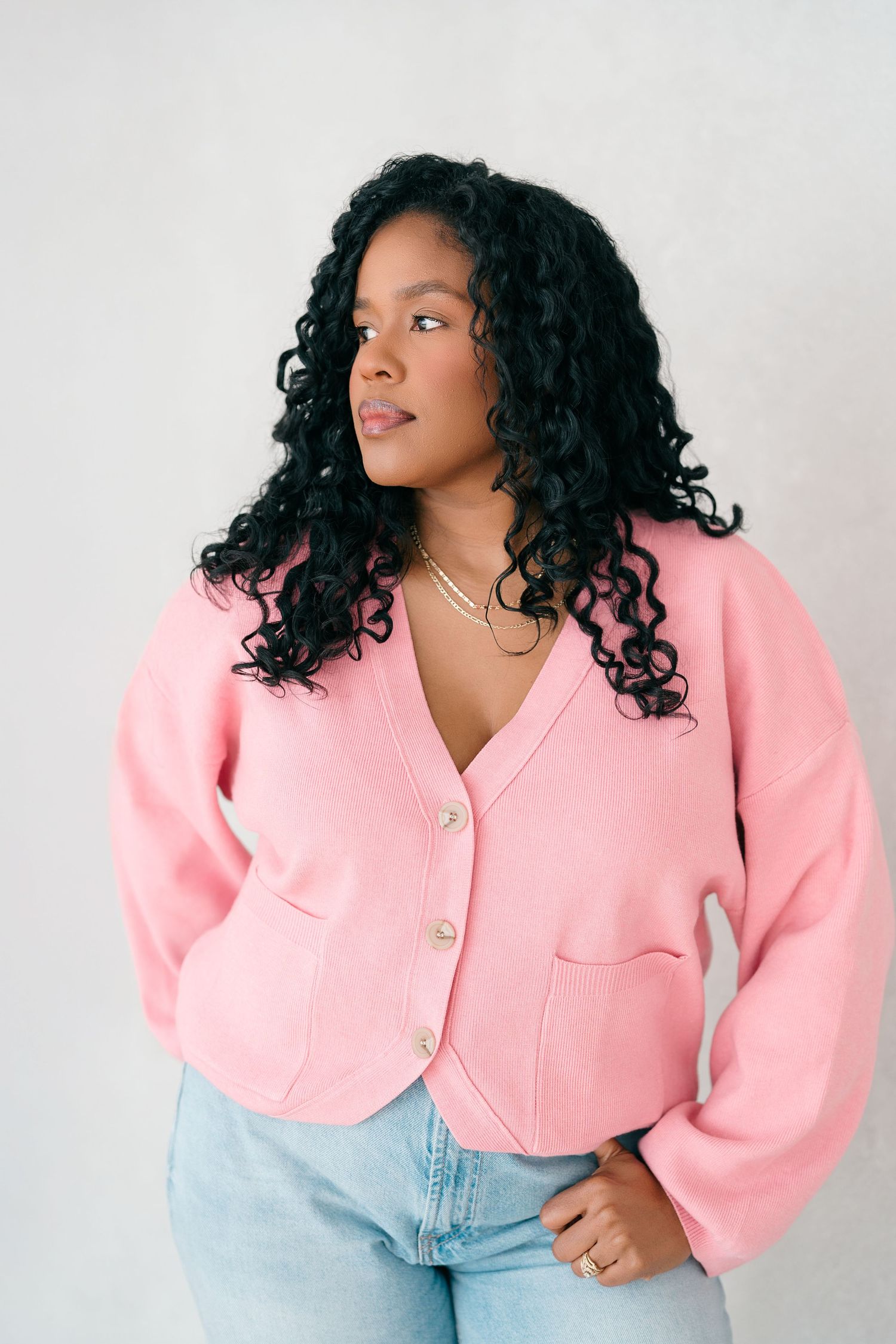 Woman in pink button-up cardigan and jeans with curly hair posing against white background.