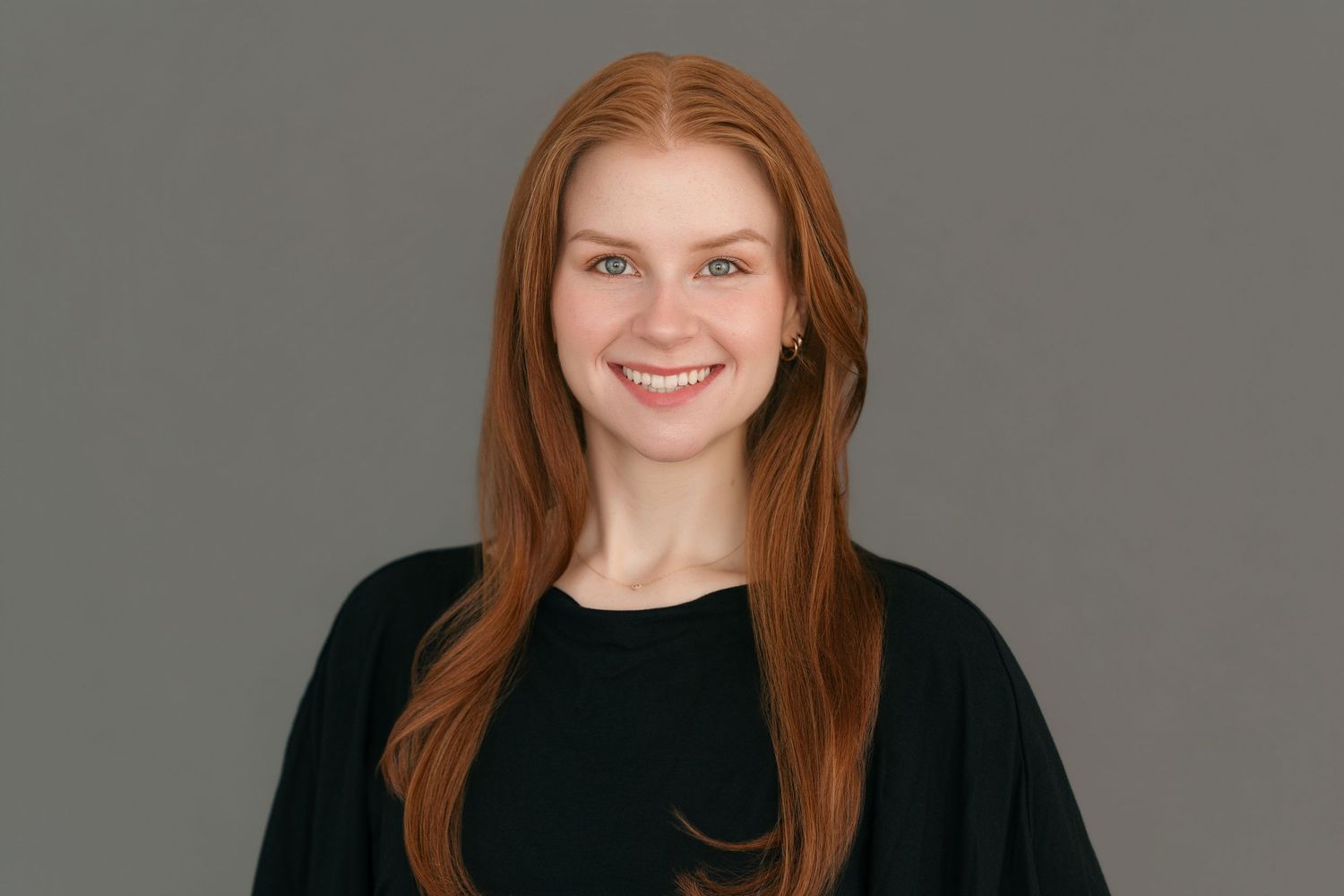 Smiling young woman with long red hair wearing a black top against a gray background.