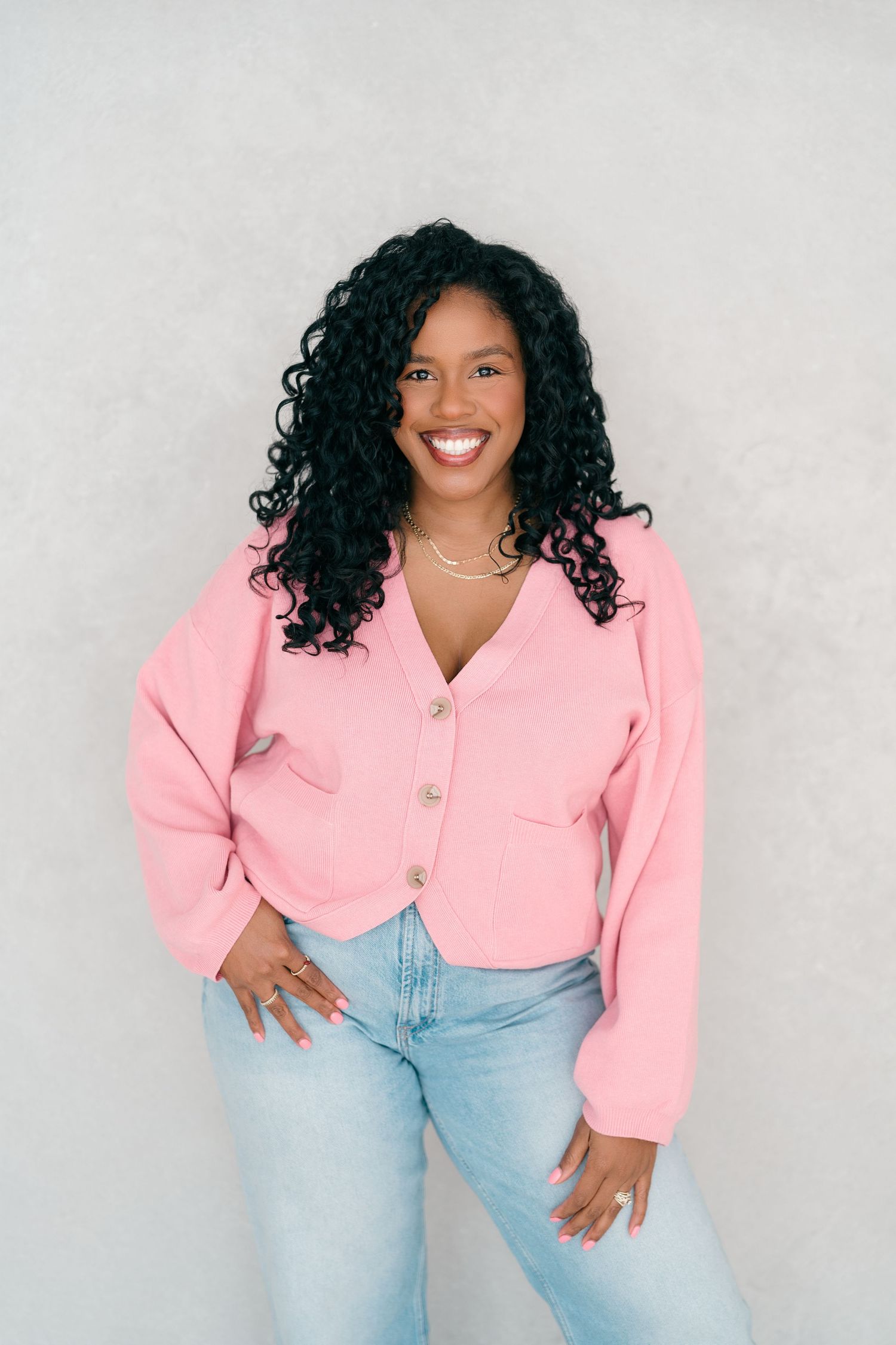 Smiling woman in pink cardigan and jeans poses confidently against a light gray background.