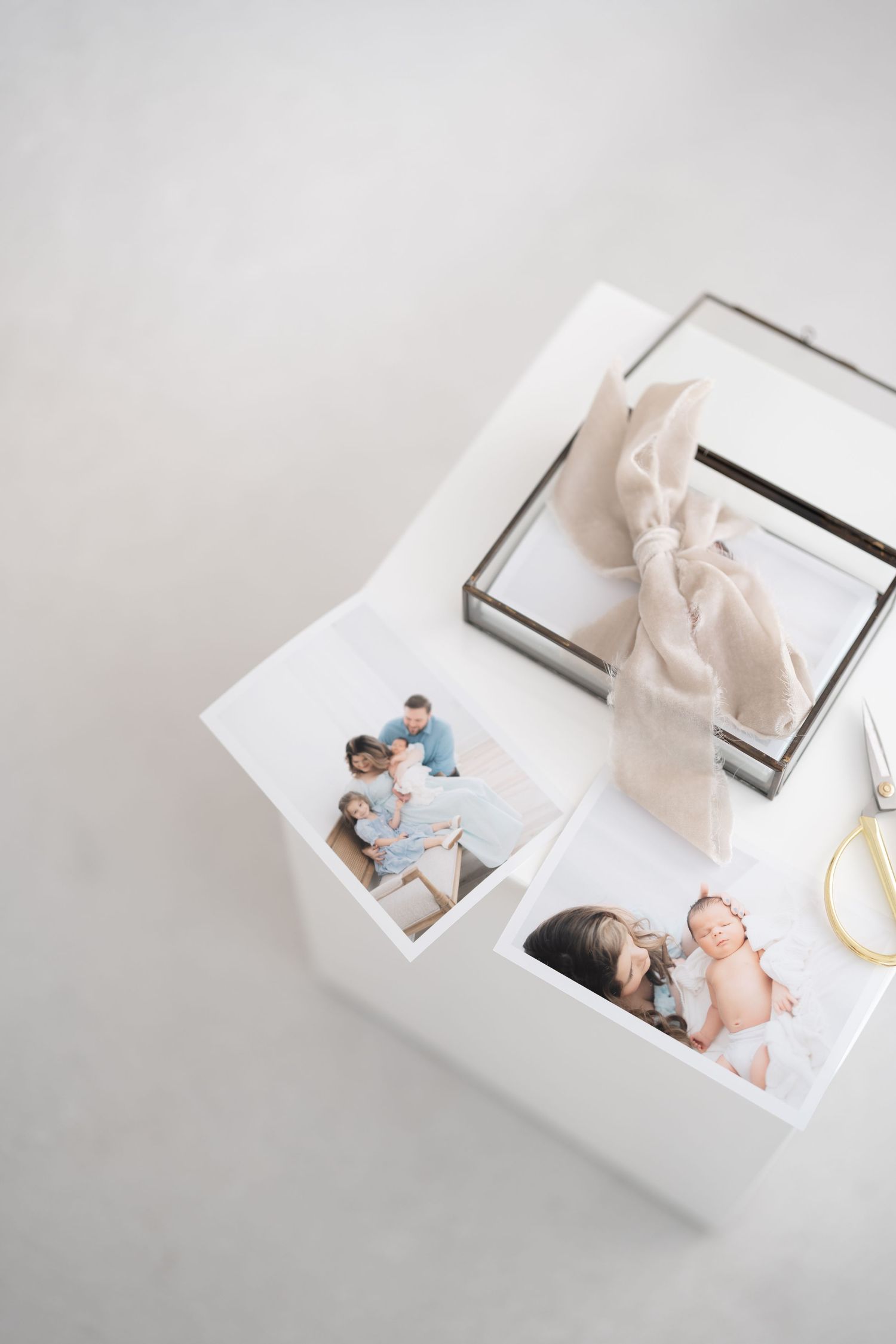 White photo box with family and newborn prints, glass keepsake box with linen ribbon, and gold scissors on light background.