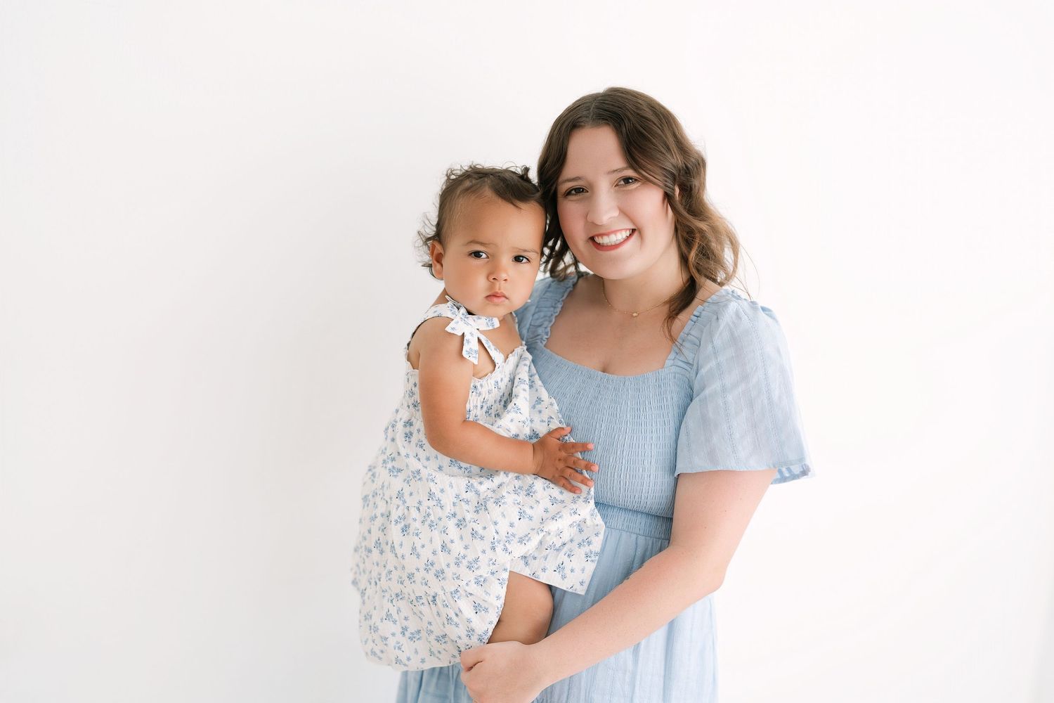 A smiling woman in a blue dress holds a cute baby girl in a floral outfit against a white background.