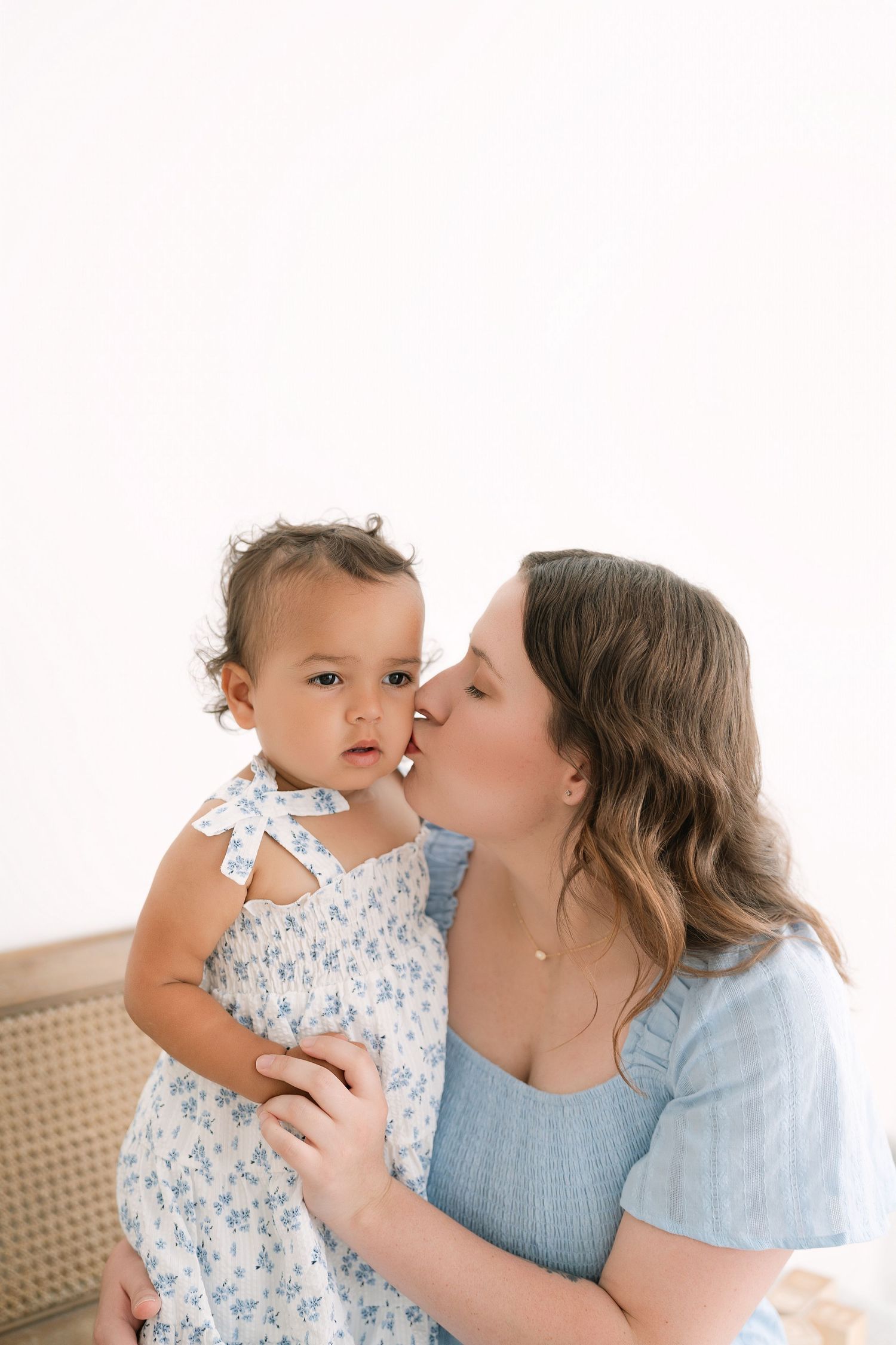 A woman in a blue dress kisses a baby girl in a floral dress while holding her against a white background.