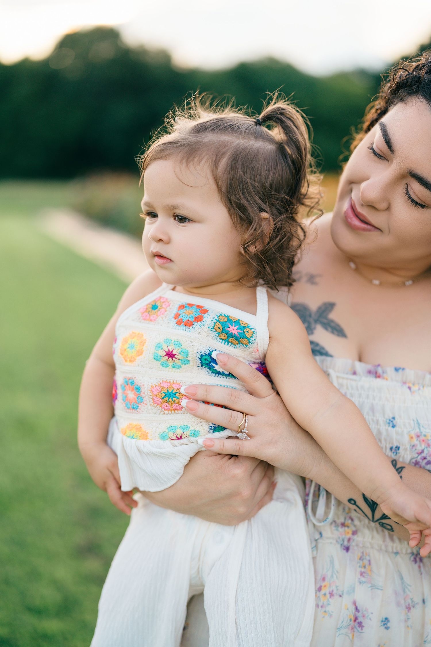 A mother holds her toddler daughter in a colorful crochet outfit outdoors in a green field.