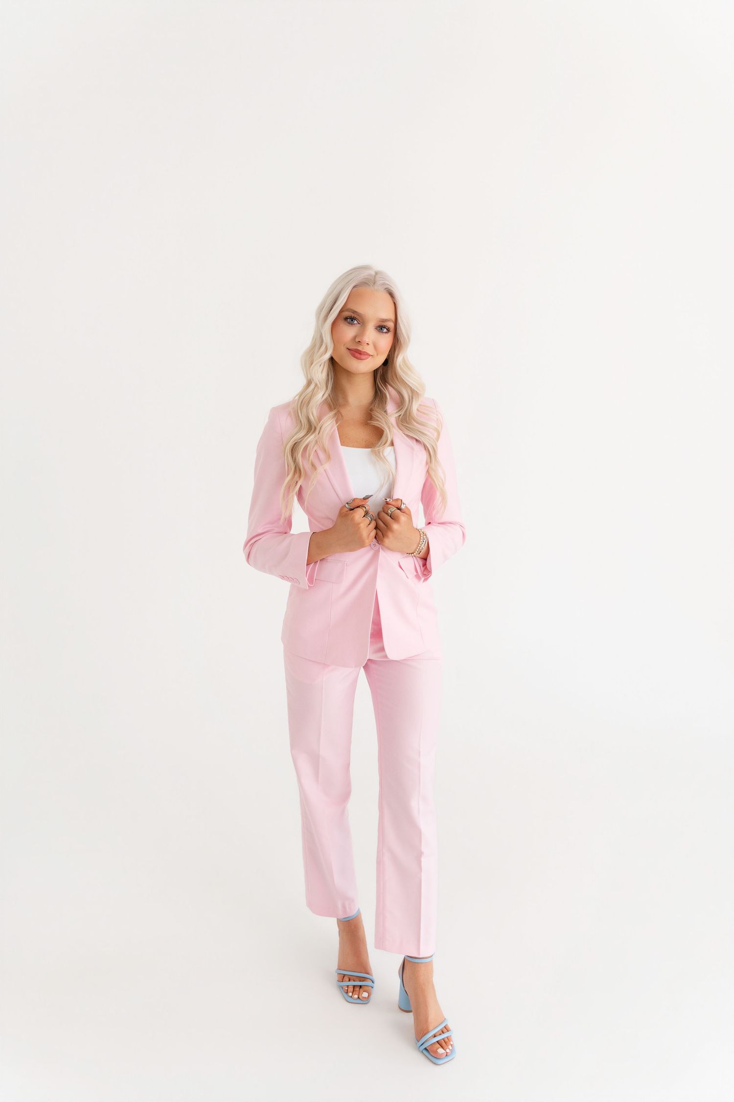 Blonde woman in a pink suit with white top and blue heels standing on white background.
