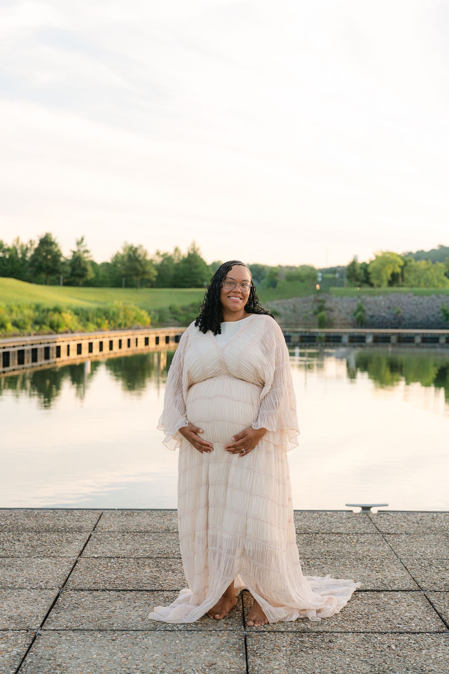 Smiling pregnant woman in flowing white maternity gown standing by a serene pond at golden hour.