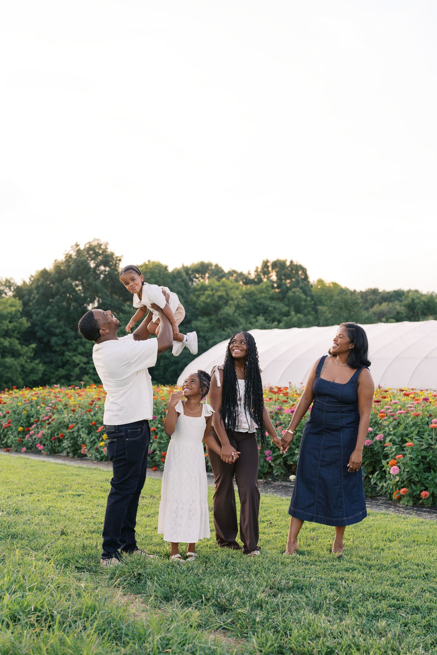 A joyful Black family plays outdoors near a flower farm, with a father lifting a baby while children and mother smile.
