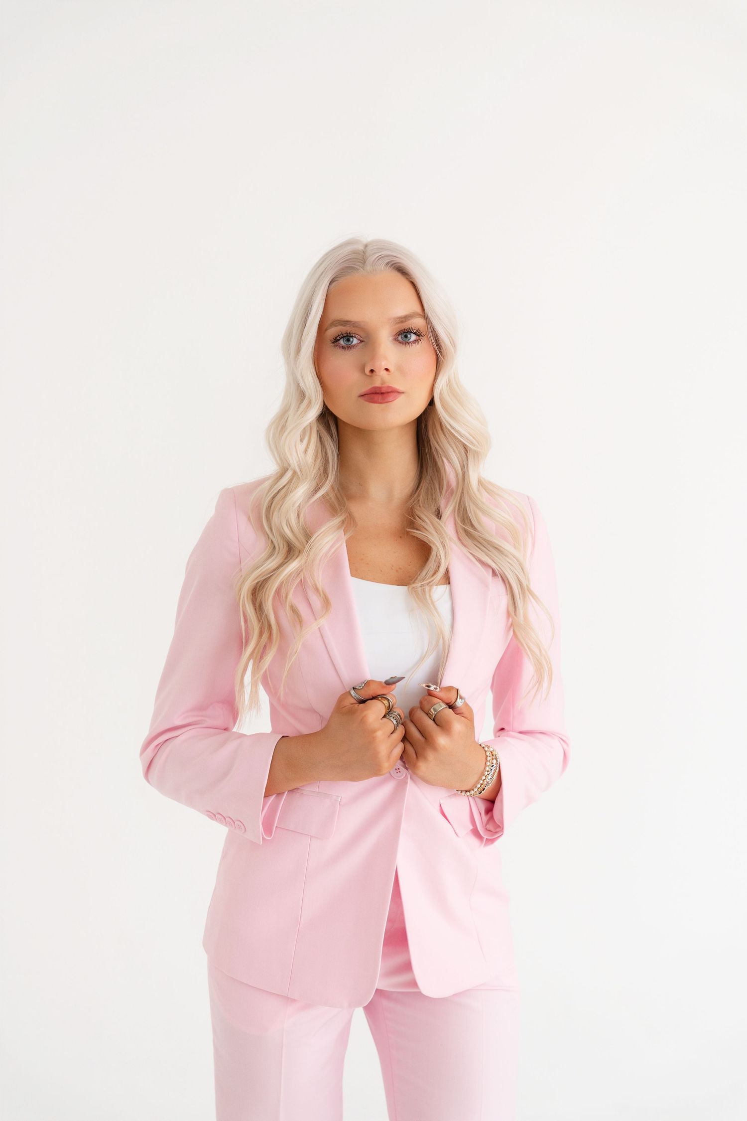 Young woman wearing a pink blazer suit with white top, long blonde wavy hair, posing on white background.