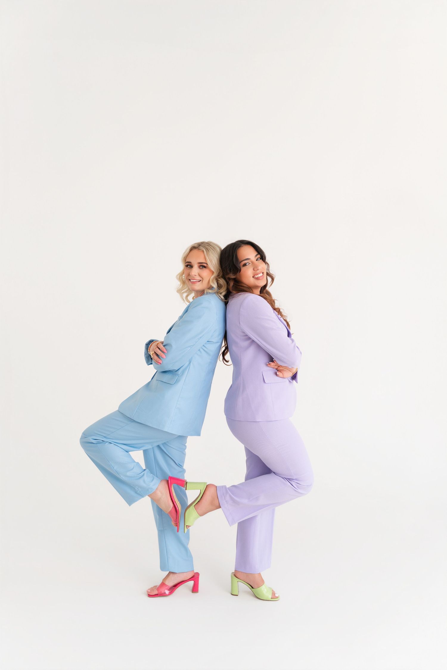 Two women in matching pastel suits pose back-to-back, wearing colorful heels on a white background.