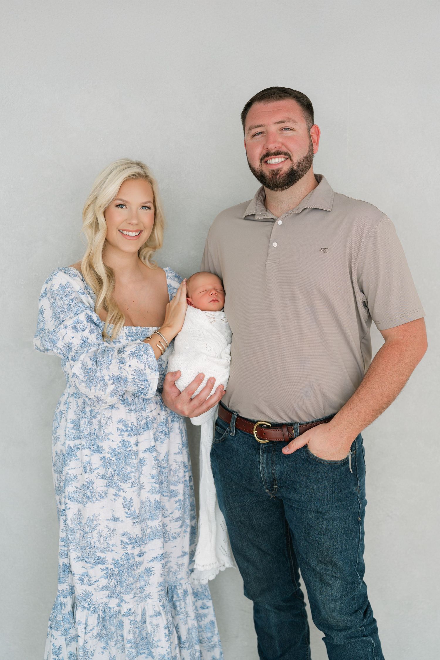 Smiling couple in studio holding swaddled newborn baby, woman in blue floral dress, man in tan polo and jeans.