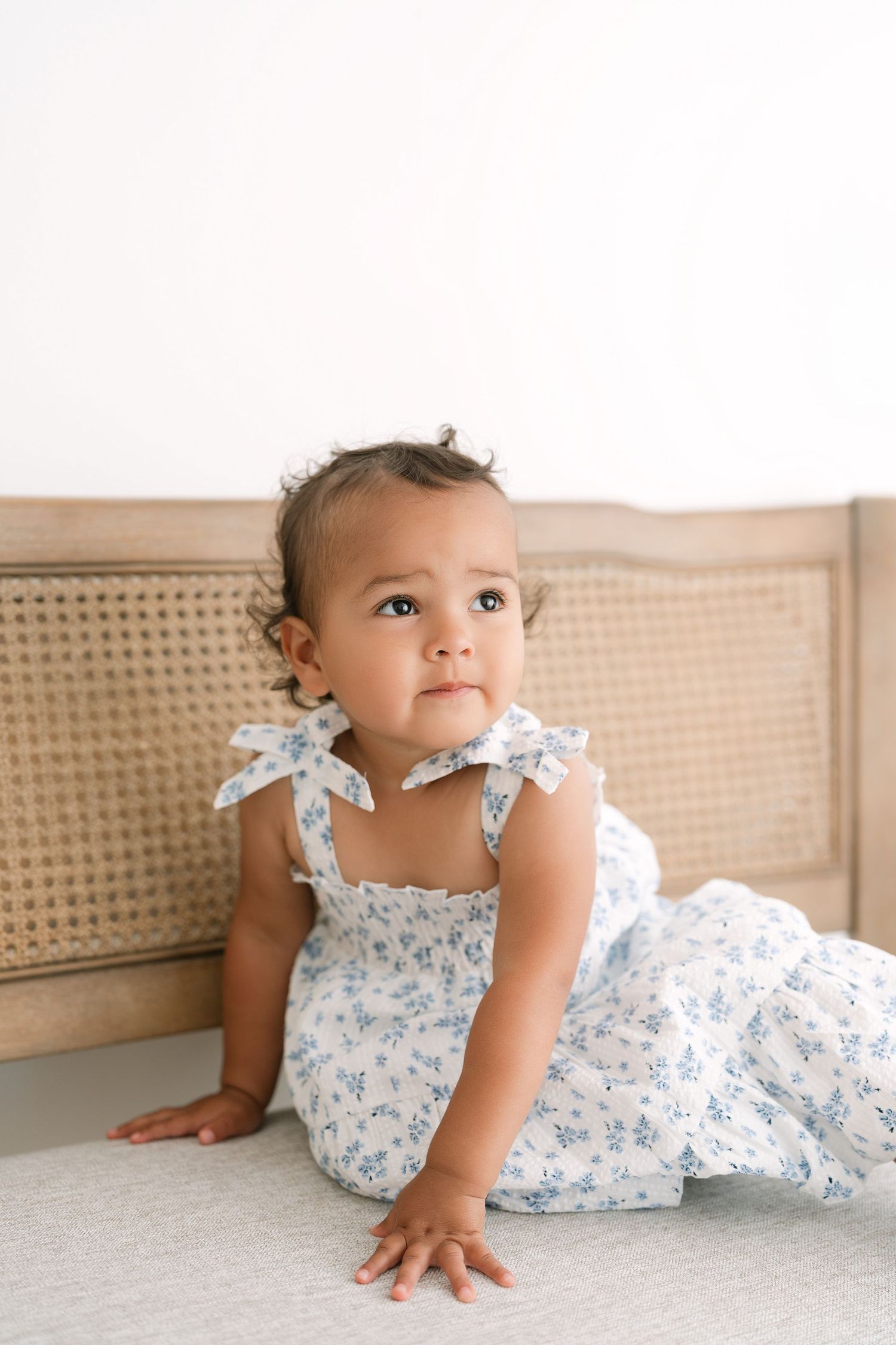 Adorable baby girl in white floral dress with bow straps sitting on bed leaning against rattan headboard.