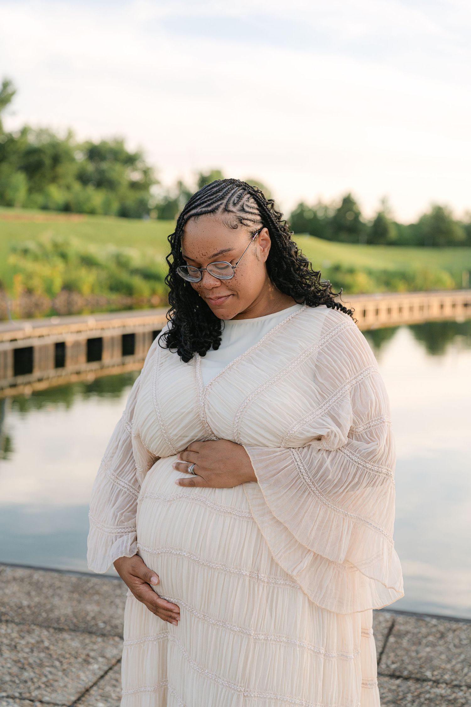 Pregnant woman in white dress holding belly by a serene pond, wearing glasses with braided curly hair.