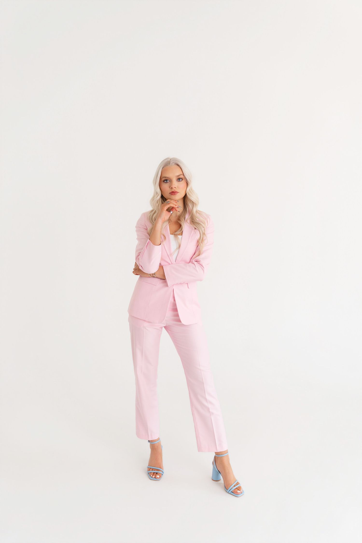 Woman posing in a pink blazer and matching trousers with blue heels on white background.