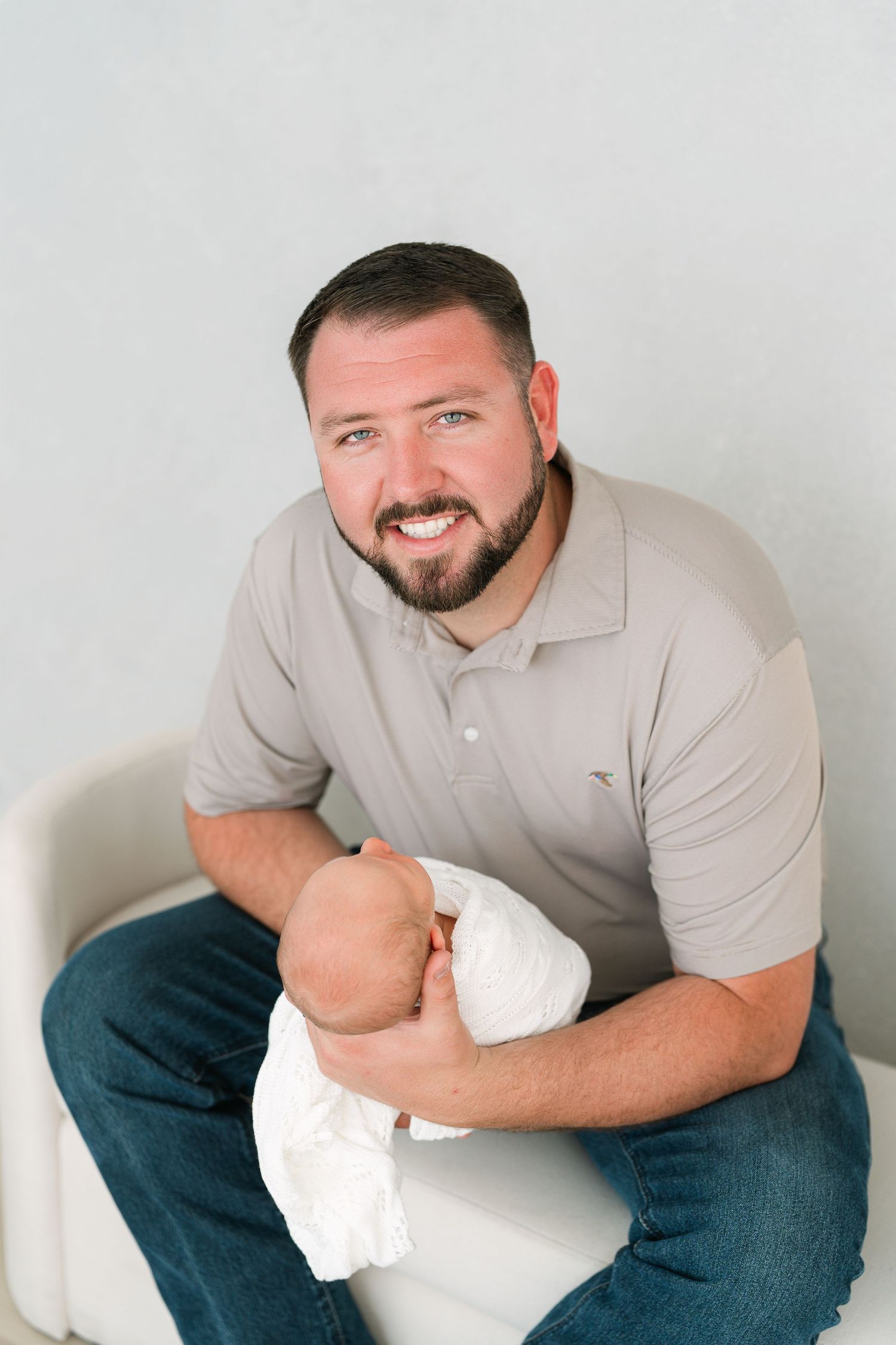 Smiling bearded man in gray polo shirt holding a swaddled newborn baby while sitting on a white chair.
