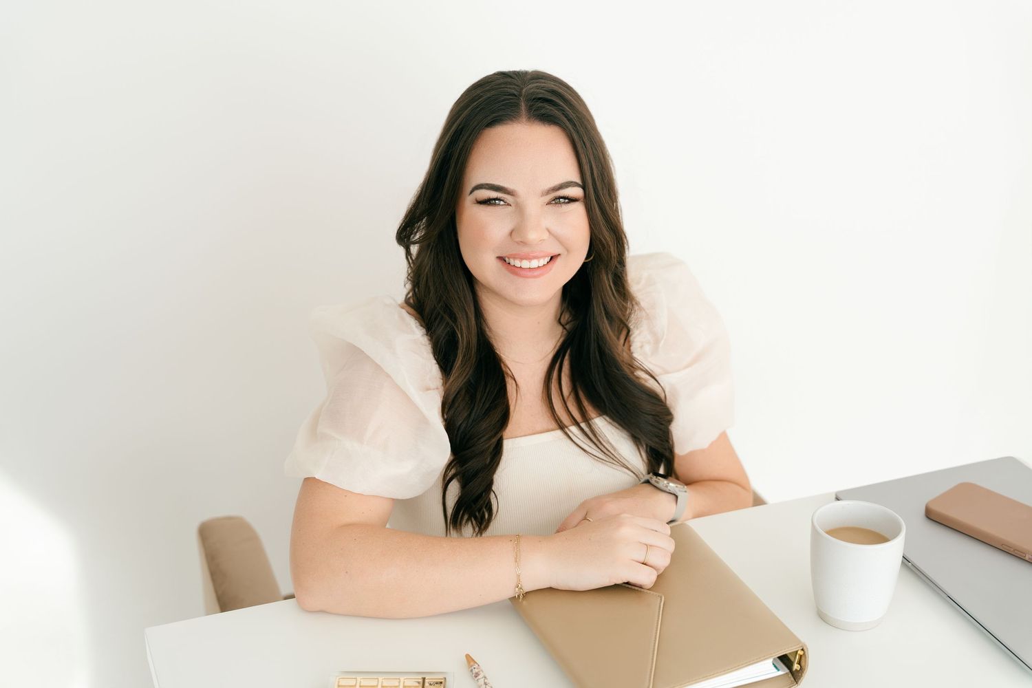 Smiling brunette woman in white top sitting at desk with notebook and coffee cup in bright white office.
