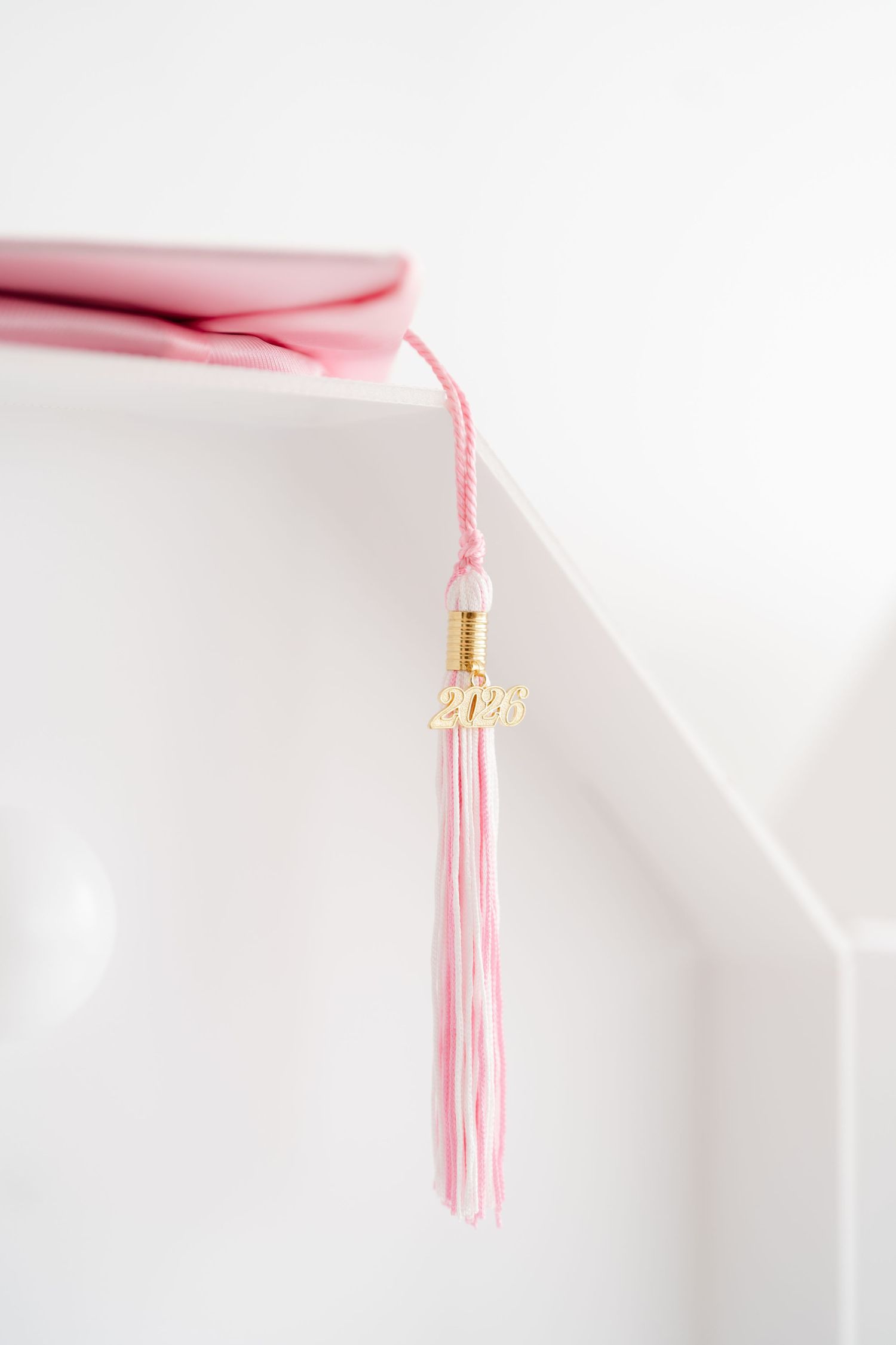 Pink graduation cap with gold 2026 tassel charm resting on white surface, celebrating academic achievement.