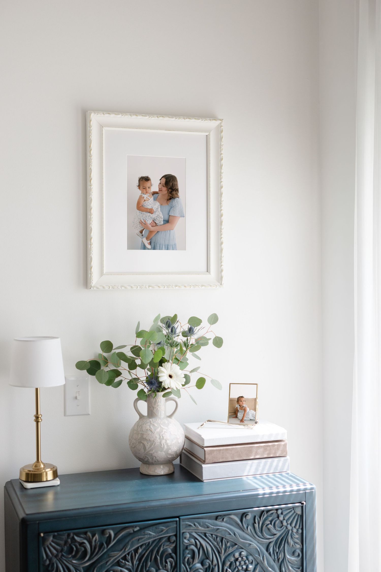 A teal carved dresser styled with a ceramic vase, eucalyptus, white flowers, stacked books, and a framed family photo on the wall.