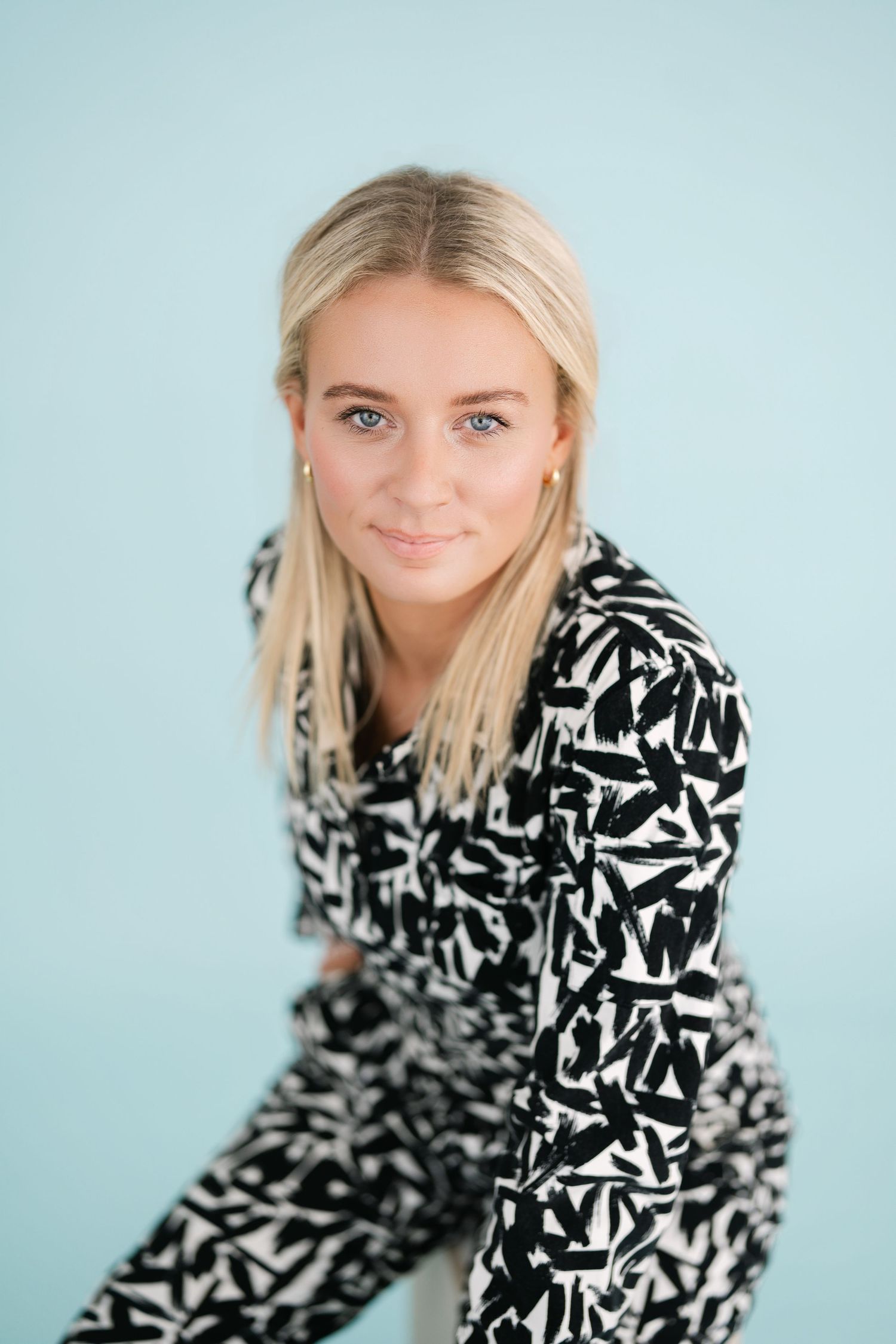 Young blonde woman smiling in black and white patterned outfit against light blue background.