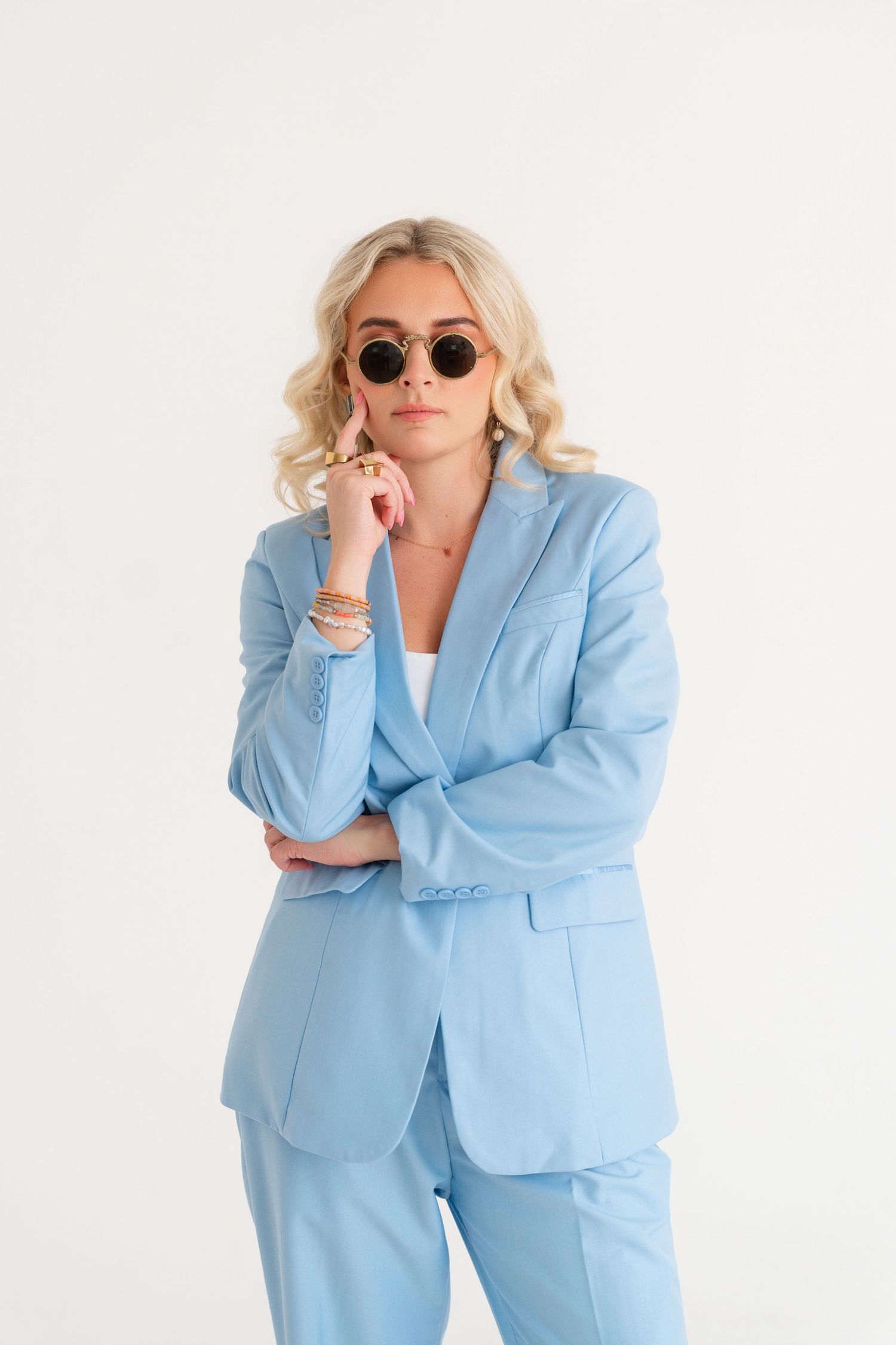Blonde woman in light blue pantsuit and round sunglasses posing against white background.