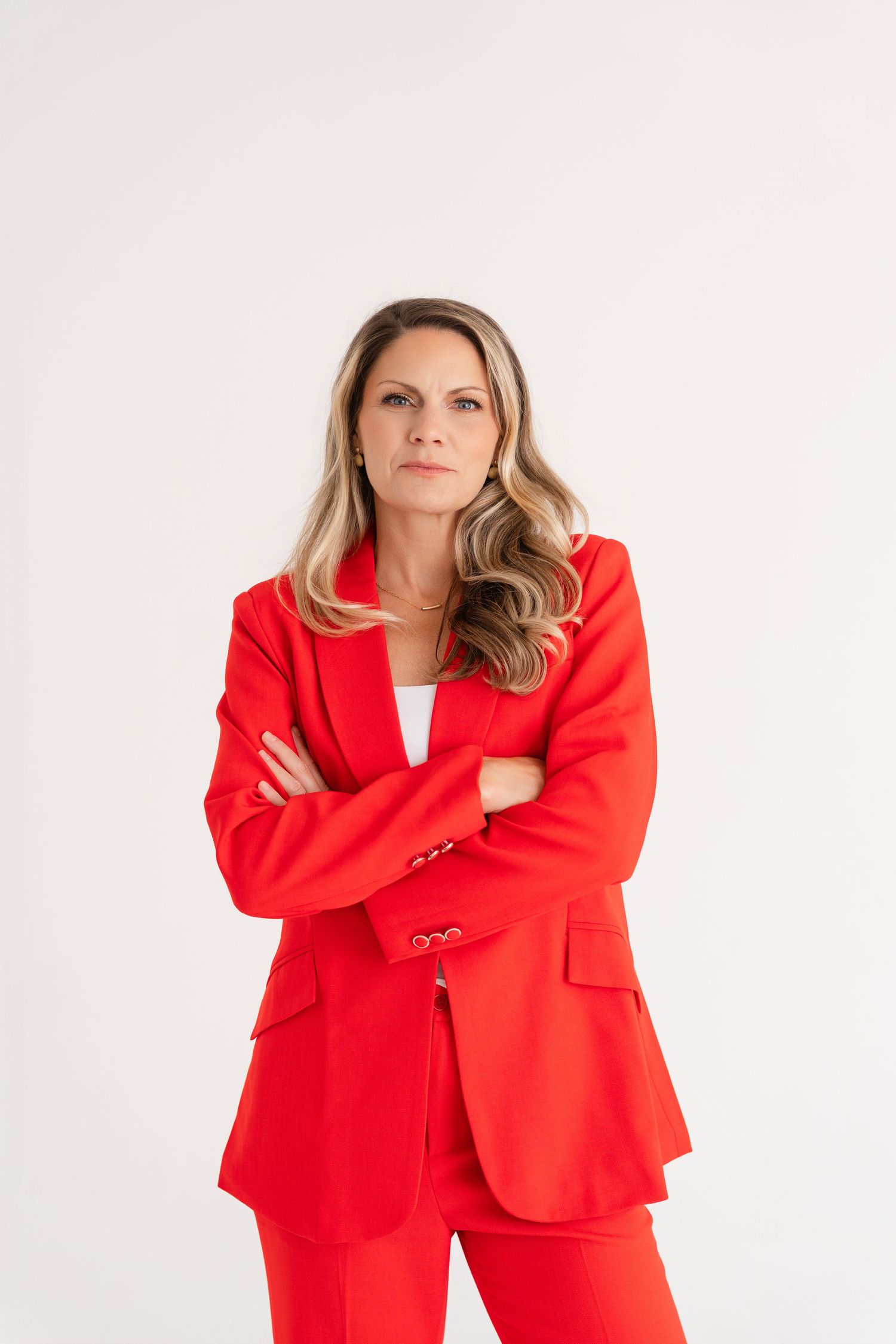 A confident woman in a bold red suit stands with arms crossed against a white background.