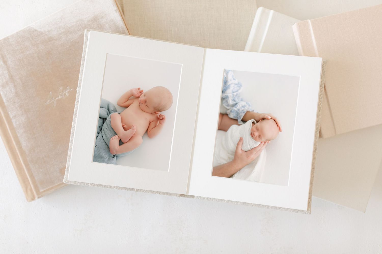 Open photo album displaying two newborn baby portraits in white matted frames, with linen-covered books in the background.