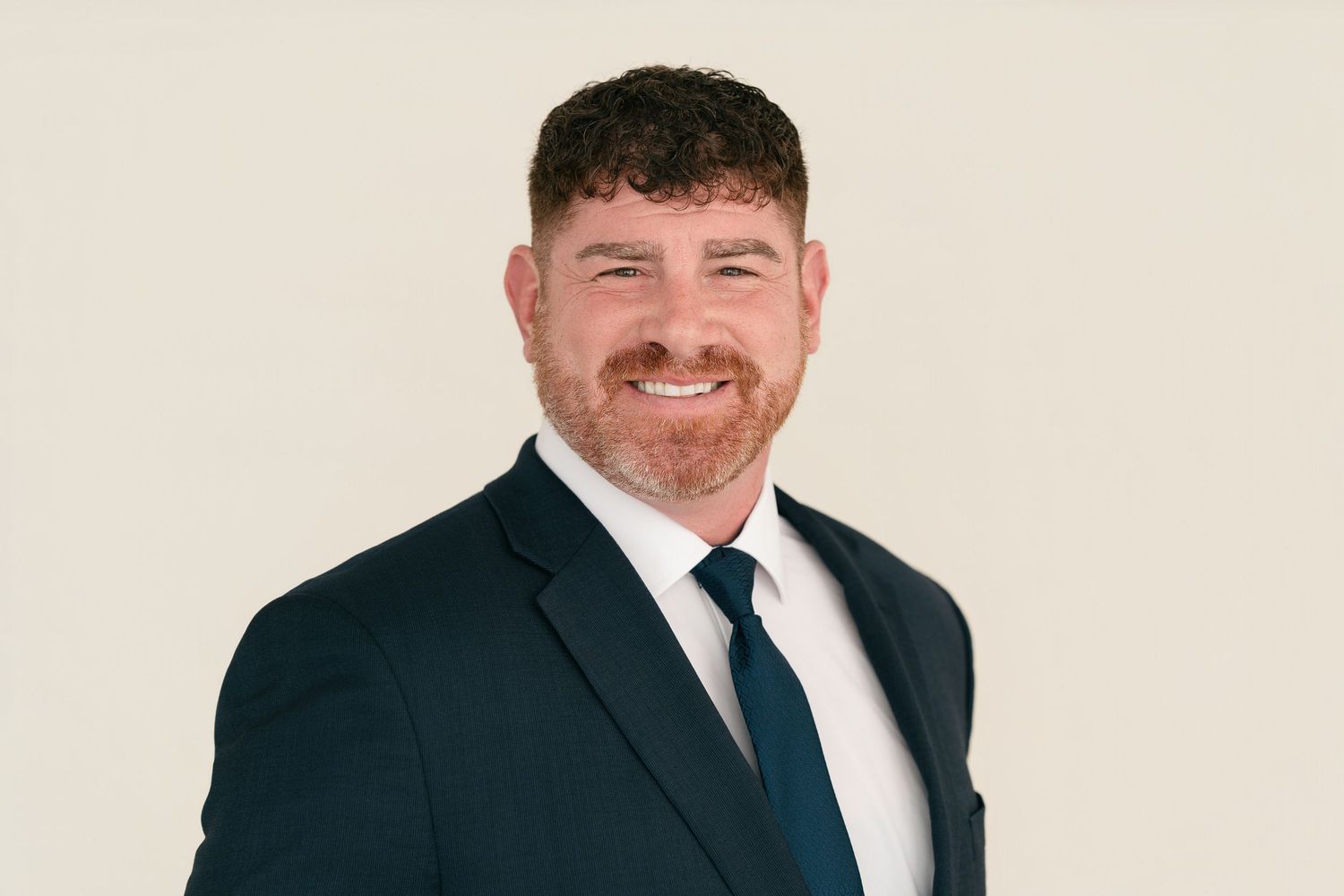 Professional man in dark suit and tie smiling confidently against a neutral background.