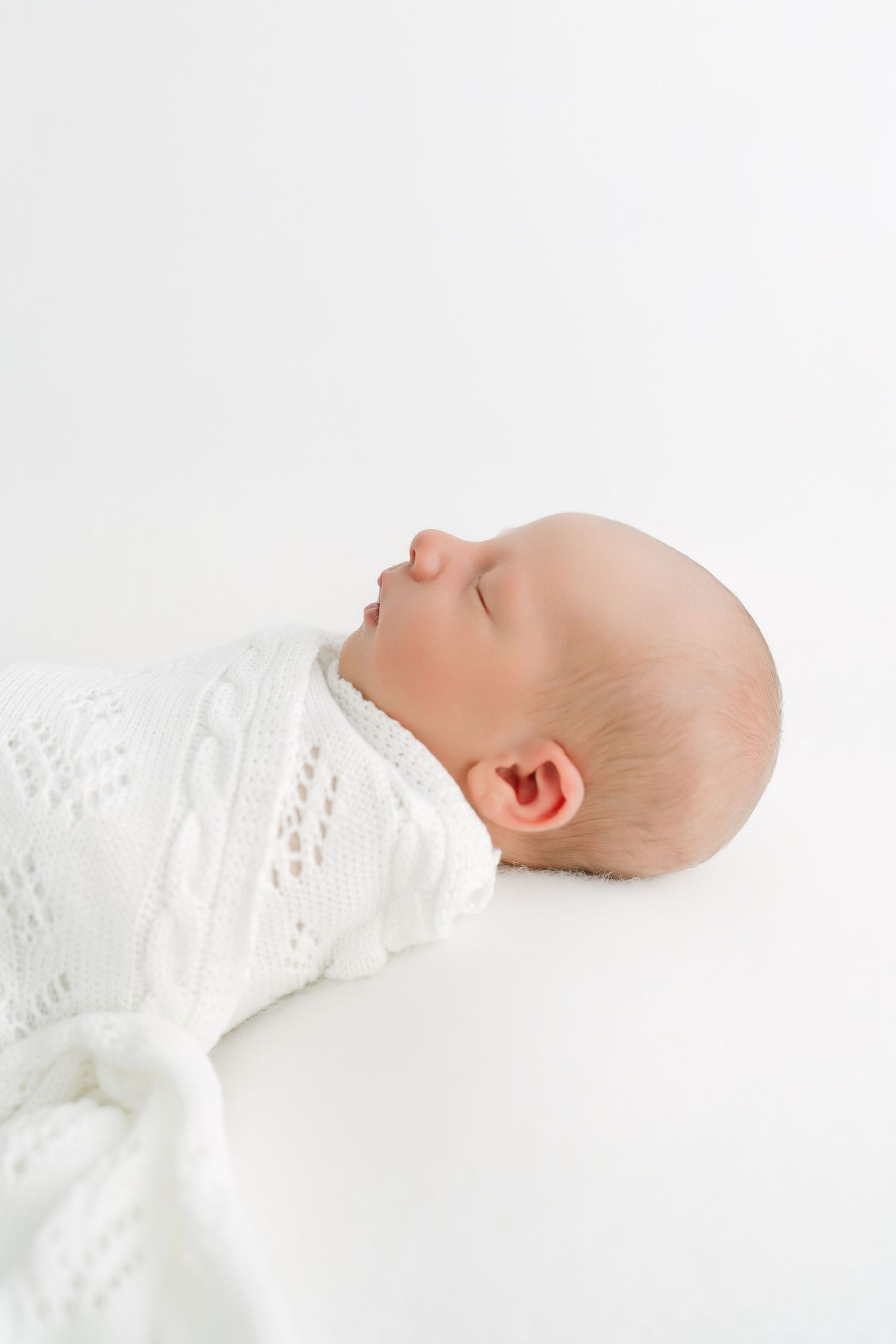 Peaceful newborn baby swaddled in white knit blanket sleeping on white background, side profile view.