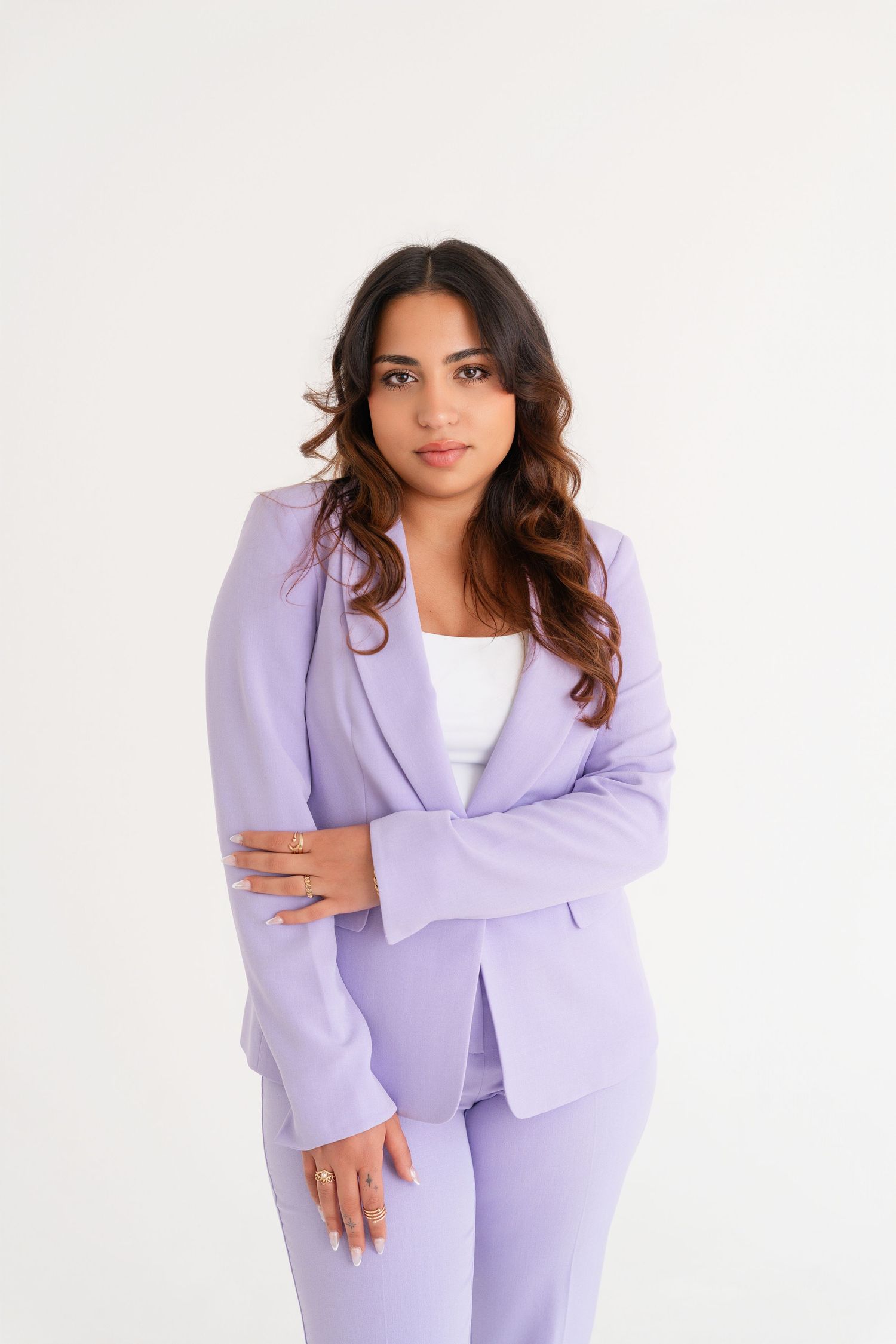 Woman in lavender pantsuit with wavy hair poses confidently against white background.