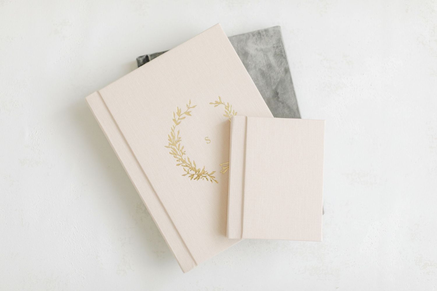Elegant cream linen photo album with gold floral wreath embossing beside a small notebook on white background.