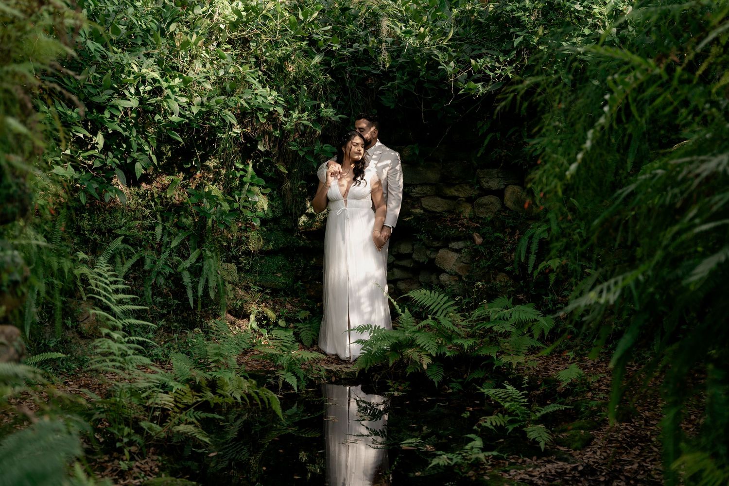 Enchanted forest couple photoshoot. A couple stands on a wooden bridge surrounded by lush green ferns in a magical forest setting.