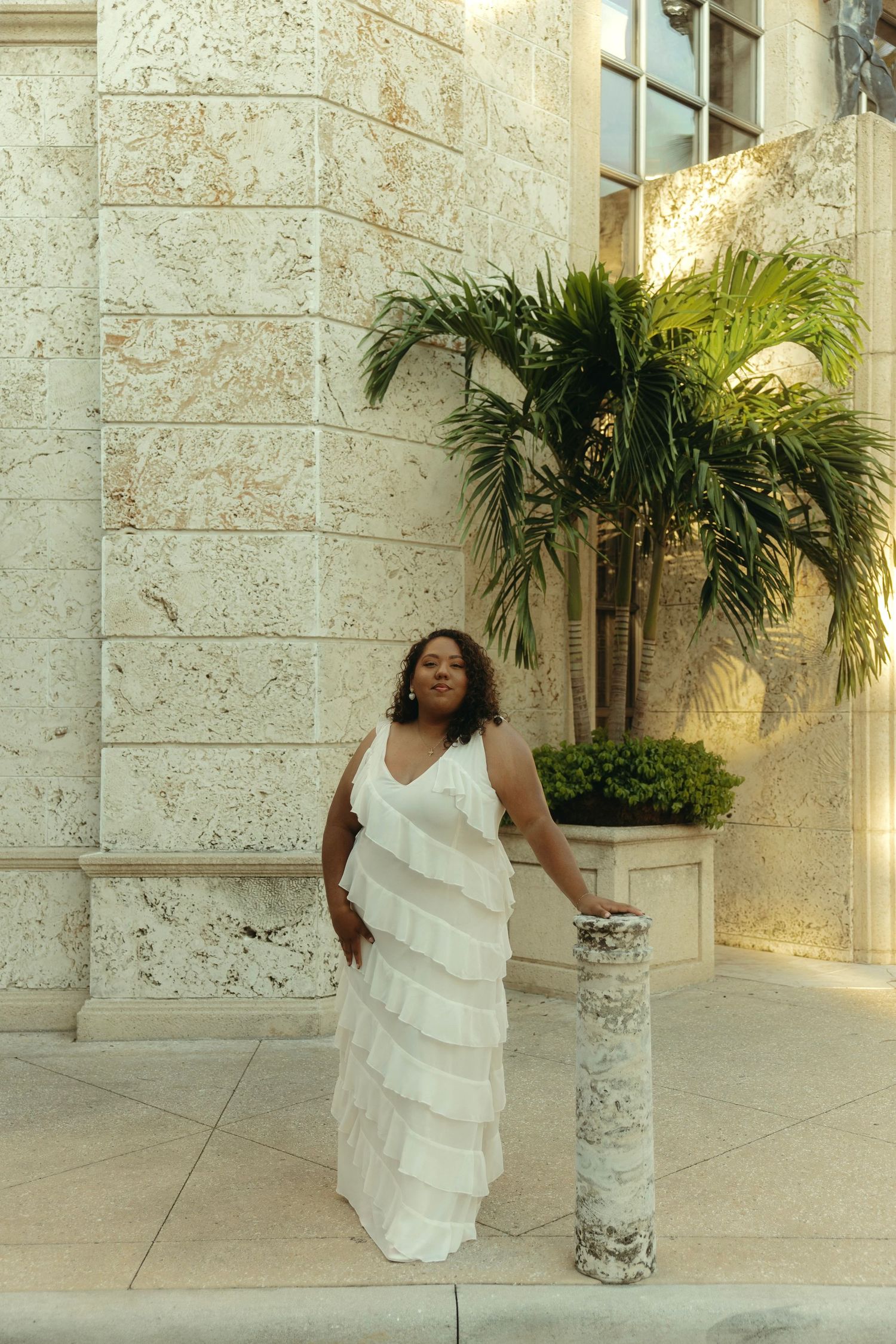 Tan woman in white flowy dress posing in front of building.