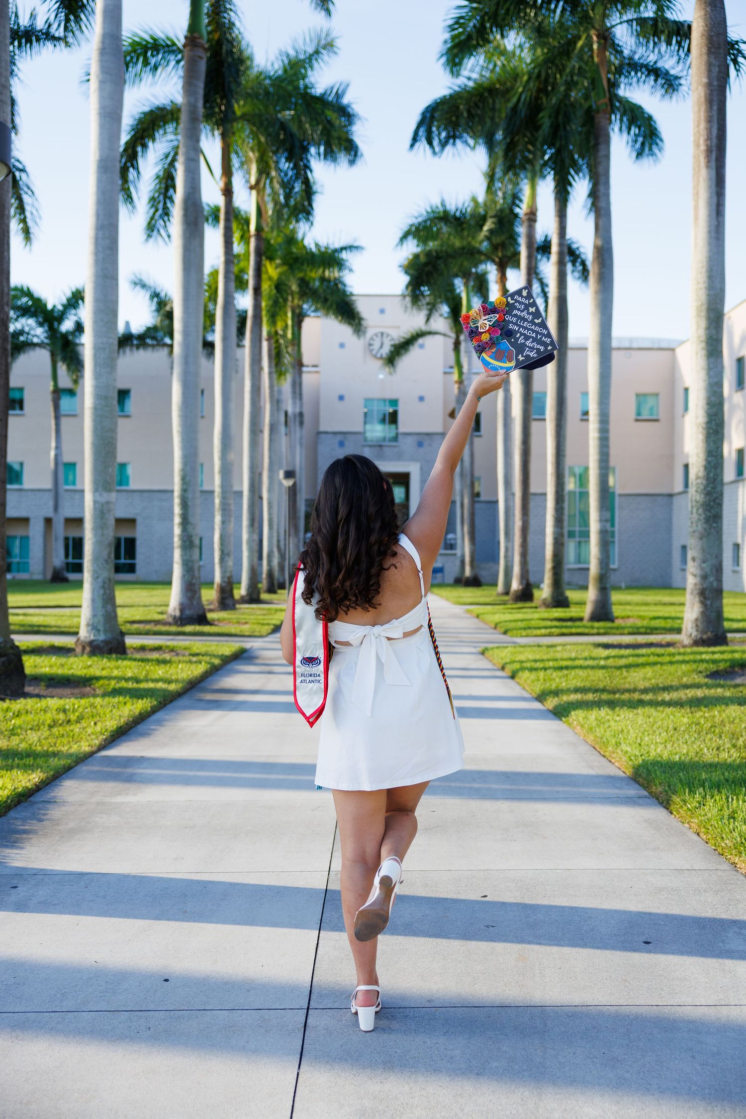 FAU graduate holding up graduation cap in front of palm trees and building.