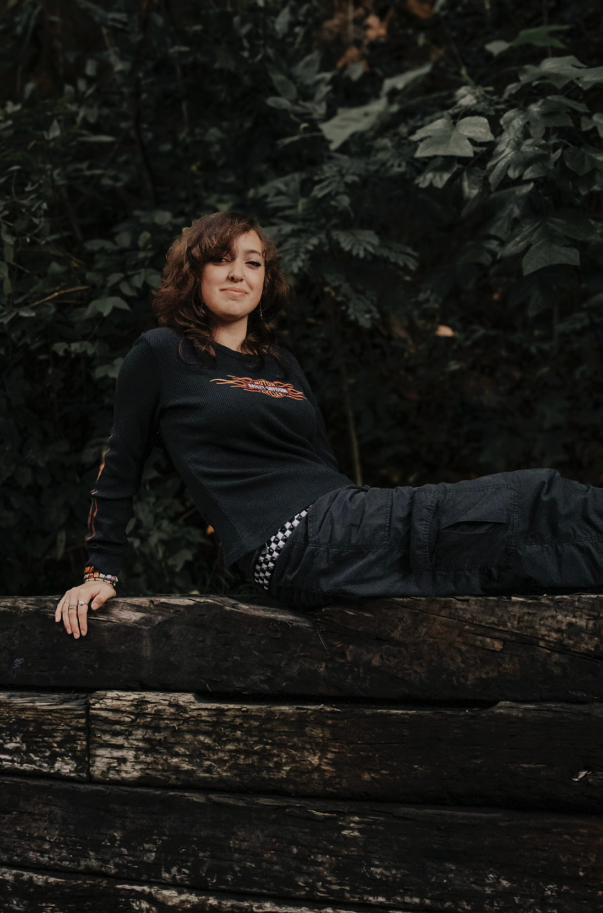 A person in black clothing reclines on a rustic wooden log against a dark foliage background.