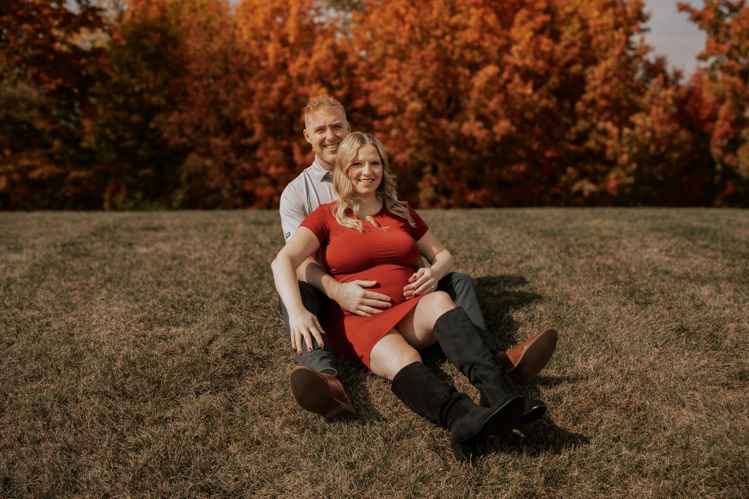 Couple sits together on autumn grass surrounded by orange and red fall foliage at sunset.