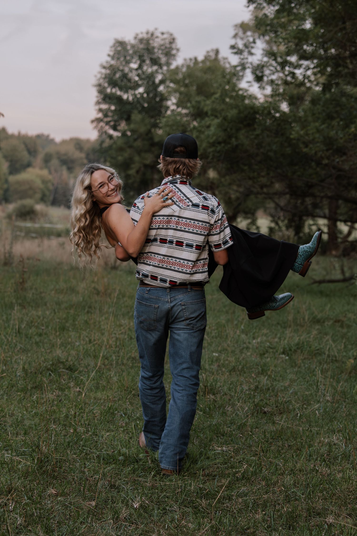 A man carries a smiling woman in a field, both wearing jeans and patterned shirts, surrounded by lush green trees.