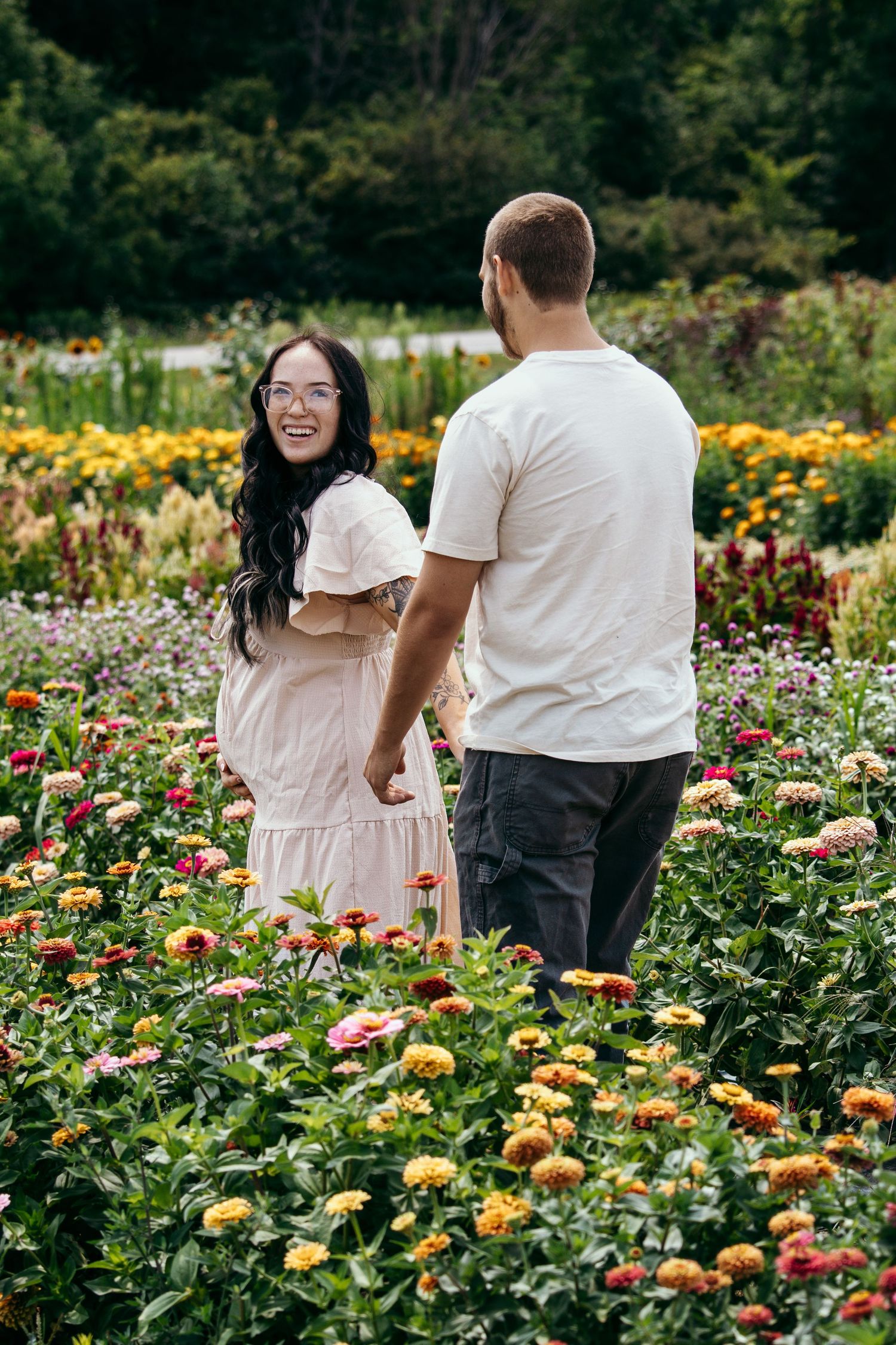 A smiling woman in a pink dress holds hands with a man while walking through a colorful flower garden.