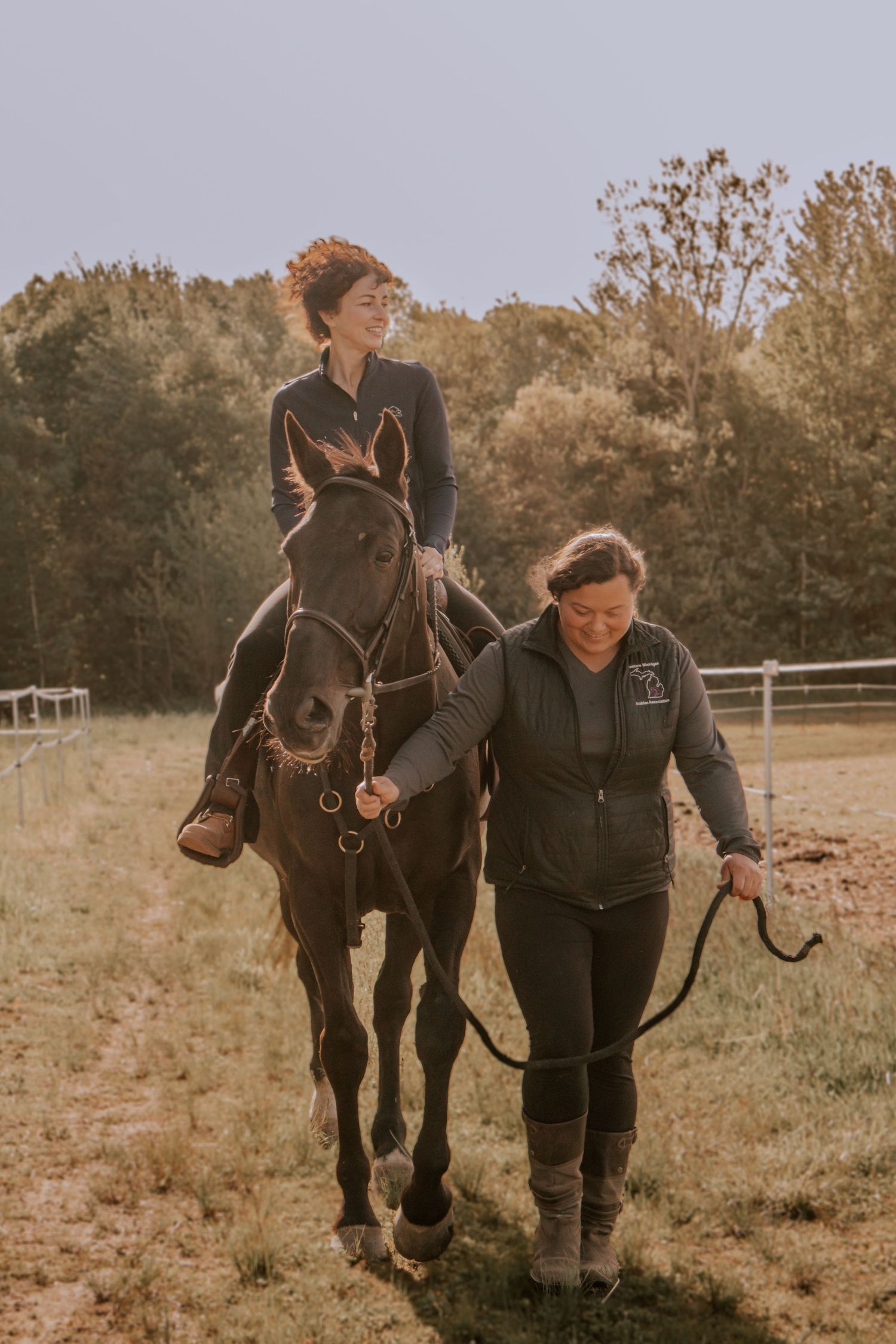 Woman riding dark horse led by instructor across sunny field with autumn trees in background.